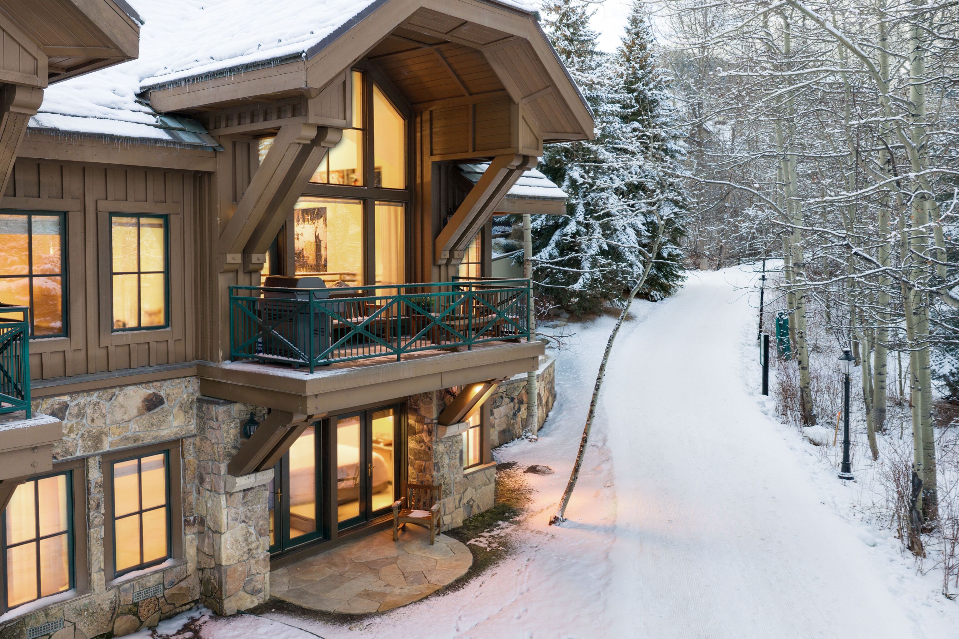 This scenic mountain-style residence showcases a beautiful blend of stone masonry and timber detailing, highlighted by oversized windows and a large balcony. The cozy, warm lighting from the interior contrasts against the tranquil, snow-covered surroundings and pine forest, offering an inviting and luxurious retreat. Ideal for winter living, the home features a private ground-level patio and direct access to a snow-packed pathway.