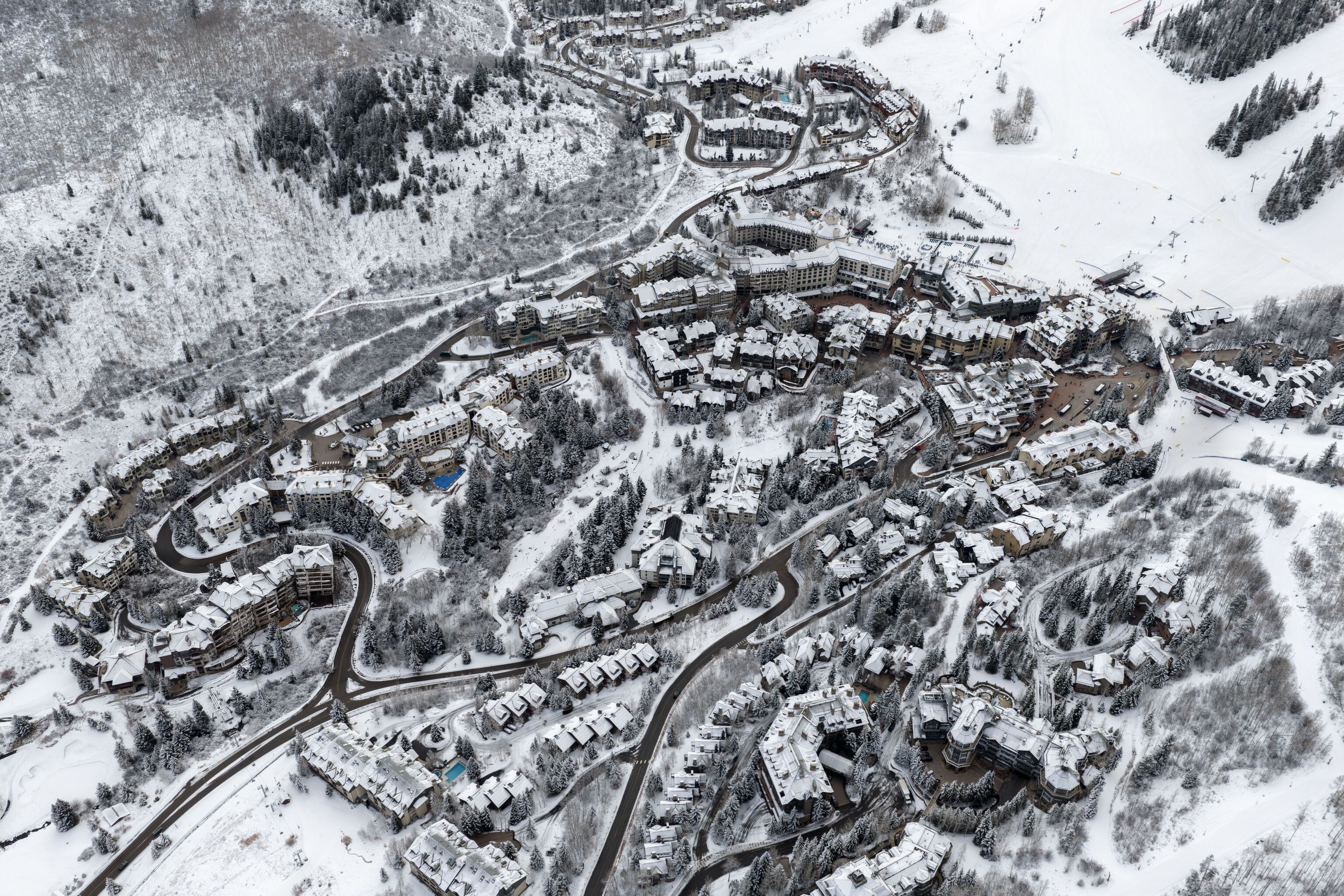 This is an expansive aerial view of a luxurious, high-altitude mountain resort community nestled in a snow-covered landscape. The image captures a dense cluster of multi-story lodge-style condominiums and townhomes connected by winding roads, set against a backdrop of heavily forested slopes and open ski runs. The scene conveys a prestigious, cold-weather retreat destination characterized by its integration into the natural alpine environment.