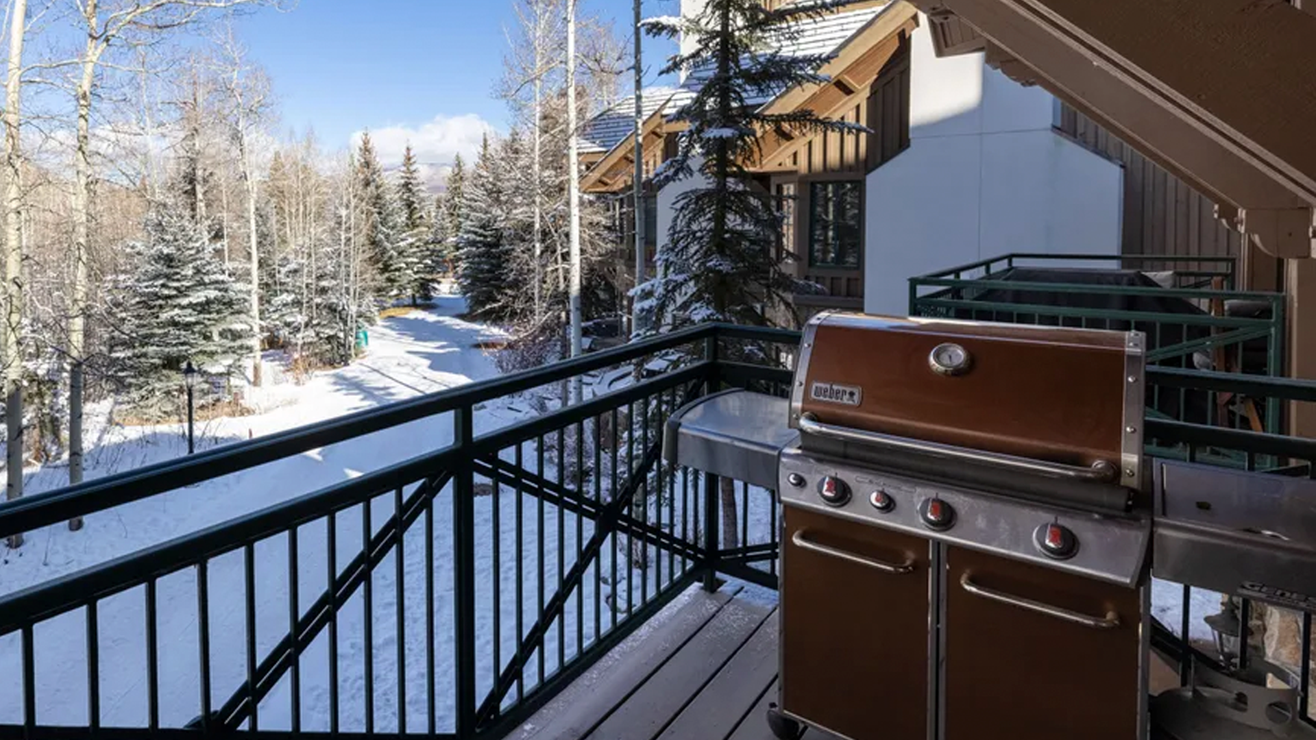 This image shows a wooden balcony overlooking a scenic, snow-covered landscape with evergreen trees and a walking path. The area is equipped with a stainless steel Weber grill, making it a perfect spot for outdoor cooking and enjoying the winter view. The setting offers a peaceful and private mountain atmosphere.