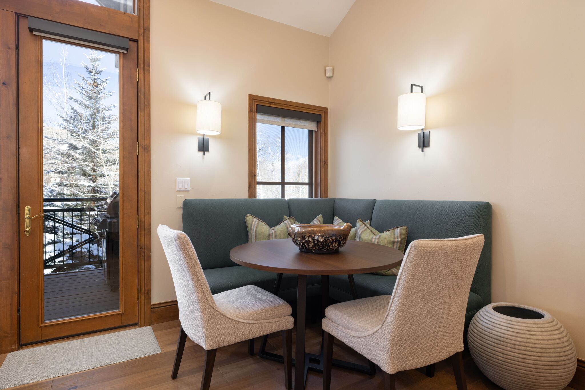 This cozy dining nook features a built-in dark teal upholstered corner banquette accented with patterned throw pillows, paired with two light-colored dining chairs and a circular wood table. The area is illuminated by two modern wall sconces and boasts natural light from an adjacent window and a glass-paned door that opens onto an outdoor deck with snowy vistas. The space combines rustic warmth with a clean, contemporary design, making it an inviting spot for casual meals.