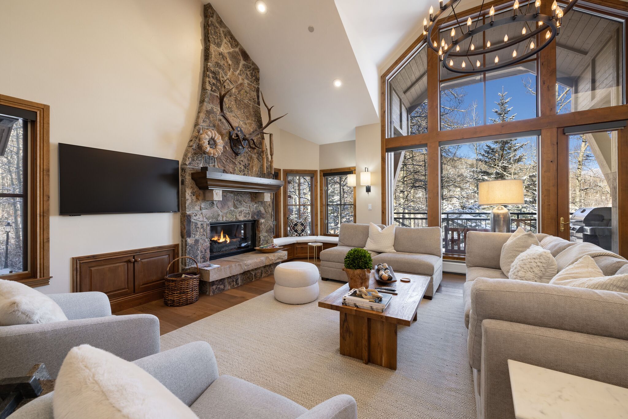 This spacious and inviting living room features a striking stone floor-to-ceiling fireplace as its focal point, complemented by soaring vaulted ceilings and large windows that offer expansive views of the snowy outdoors. The room is decorated in a refined rustic style with plush light-toned seating, wood trim detailing, and a modern chandelier, creating a warm and sophisticated atmosphere perfect for mountain living.