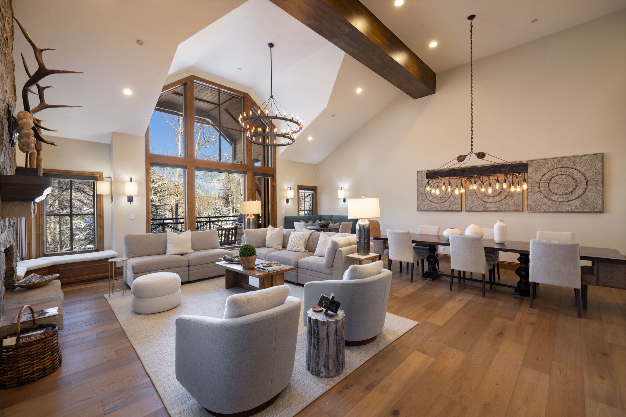 This spacious, open-concept living and dining area features vaulted ceilings with exposed wood beams and expansive windows that provide stunning natural light and scenic views. The room is decorated in a sophisticated, neutral mountain-modern palette, featuring comfortable plush seating, a rustic wood coffee table, and elegant chandelier lighting. The layout seamlessly integrates a cozy stone fireplace sitting area with a formal dining space, offering an inviting and luxurious atmosphere.