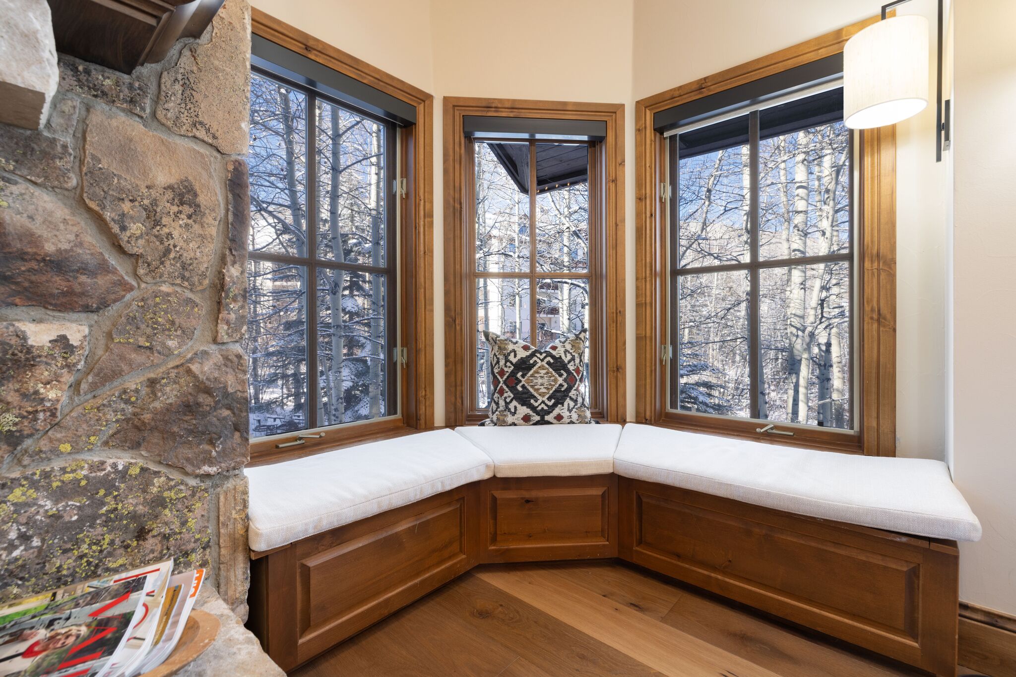 This cozy and inviting window nook features custom wooden built-in seating with plush, light-toned cushions, set against a backdrop of large windows offering serene forest views. The space is accented by rustic stone wall detailing, natural wood flooring, and a decorative patterned throw pillow, creating a warm and charming alcove for relaxing.