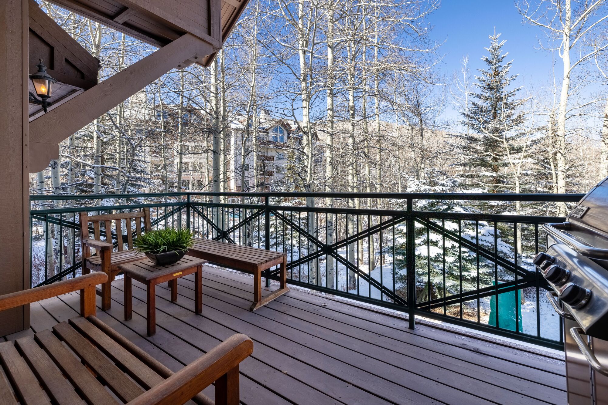 This spacious wooden balcony features outdoor seating, including benches and a small table with a potted plant, perfect for relaxing in a mountain setting. It is equipped with a stainless steel grill and provides a scenic, elevated view of snow-dusted aspen trees and adjacent hillside residences. The covered deck offers a cozy, private atmosphere ideal for enjoying the crisp winter landscape.