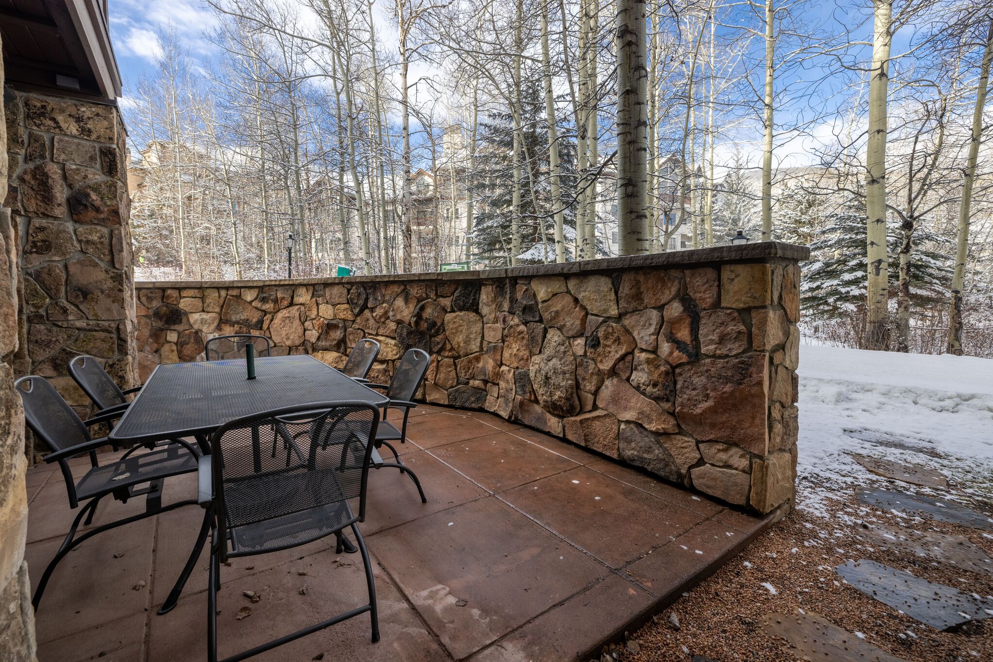 This outdoor patio features a rustic stone wall barrier that provides privacy while maintaining a natural aesthetic. It is furnished with a dark metal dining set, making it an ideal space for outdoor meals or relaxation against the backdrop of a snowy, tree-lined landscape.