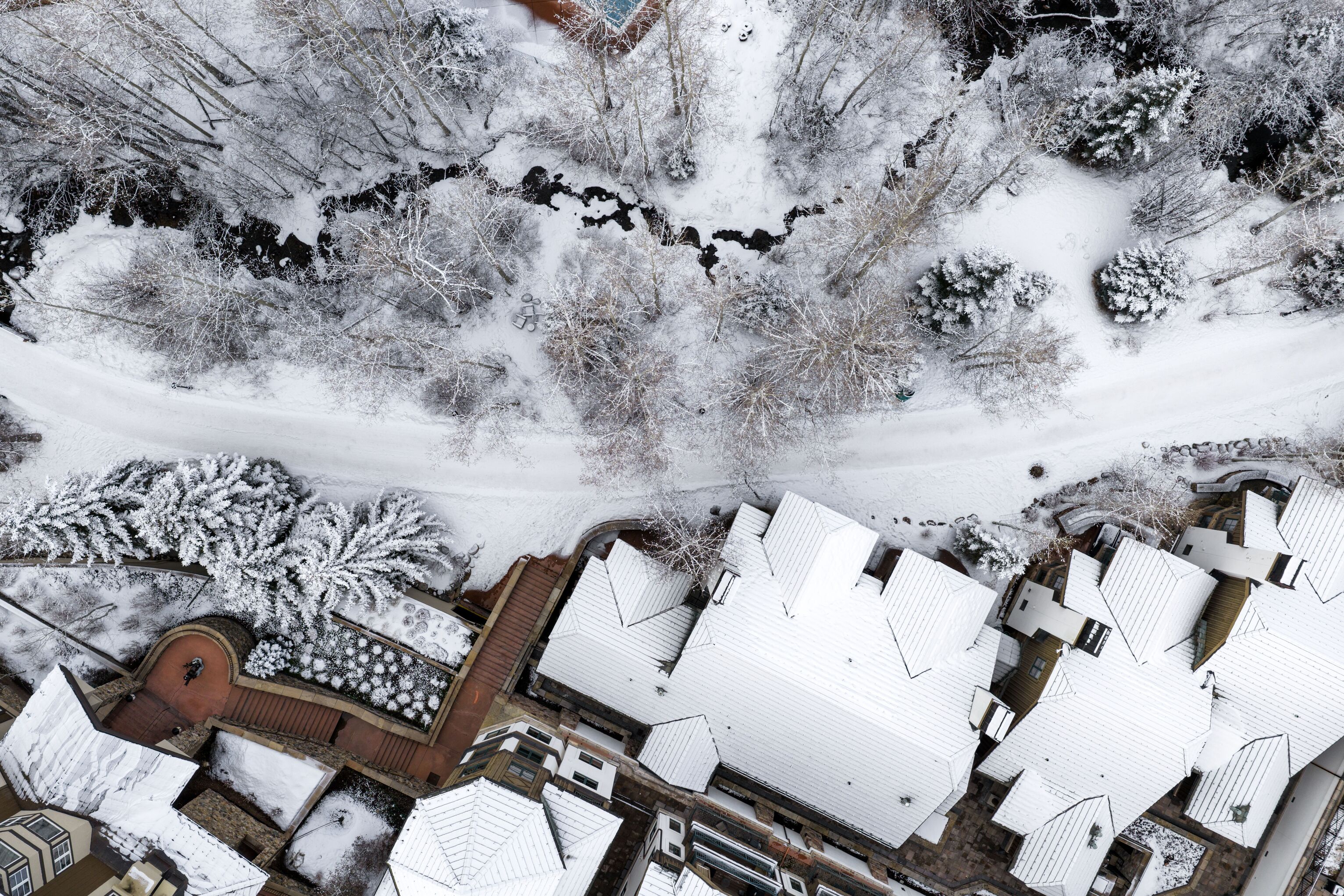 This high-altitude aerial view showcases a winter setting with snow-covered rooftops, winding paths, and a serene, tree-lined landscape featuring a small stream. The image captures the layout of the property, highlighting its integration with the surrounding natural mountainous topography. It provides an excellent perspective on the estate's proximity to nature and its architectural footprint.