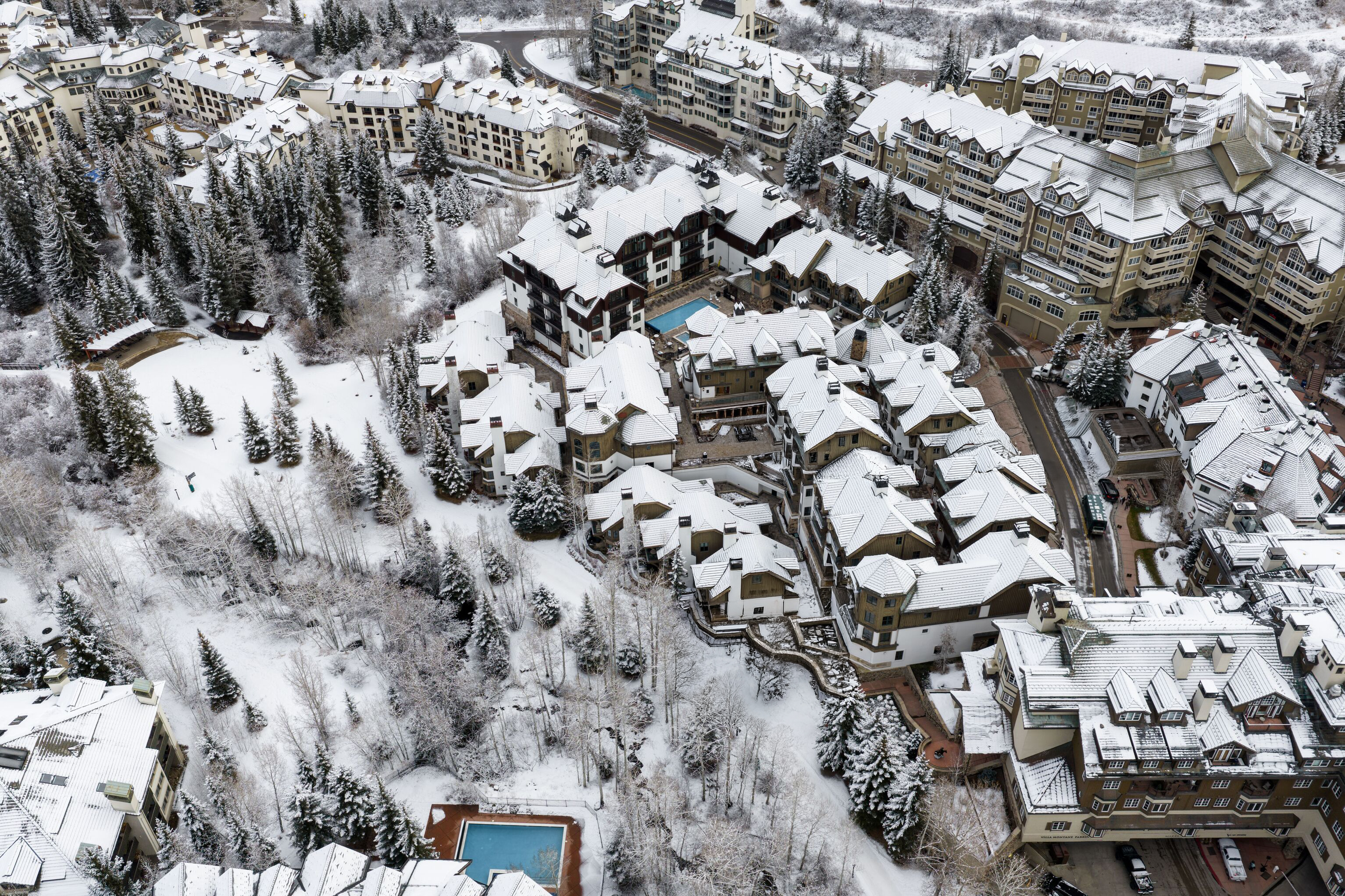 This high-altitude aerial view captures a luxury mountain resort complex nestled in a snow-covered landscape. The scene features multi-story architecture with peaked roofs finished in white snow, complemented by outdoor pools and dense pine forests. The property conveys a premium, ski-in/ski-out Alpine aesthetic, ideal for high-end winter retreats.