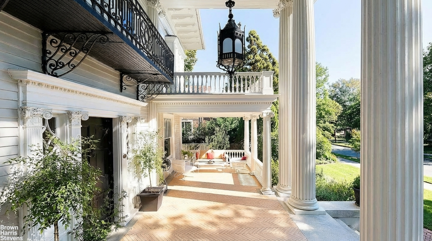 This image showcases a grand porch or patio area. Notable details include the large white columns, decorative iron detailing on the balcony above, and a brick floor adding to the exterior's upscale aesthetic. There is outdoor seating suggesting a comfortable space for relaxation and entertainment.