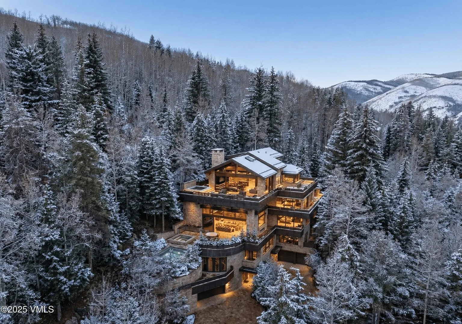 This is an aerial view of a luxury mountain home in a snowy, forested setting. The multi-level structure features stone facade, ample windows, and large decks, suggesting high-end finishes and outdoor living spaces. The integration of a pool/spa area indicates a focus on relaxation and recreation.