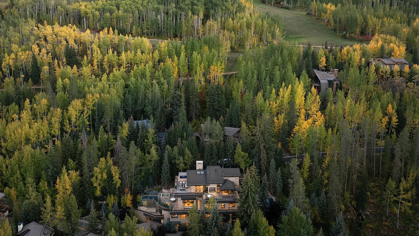 An aerial view showcases a luxurious home nestled in a dense forest with trees in their autumn colors. The multi-level house features a stone exterior, multiple balconies, and a swimming pool. The setting emphasizes privacy and natural beauty, making it an appealing retreat.