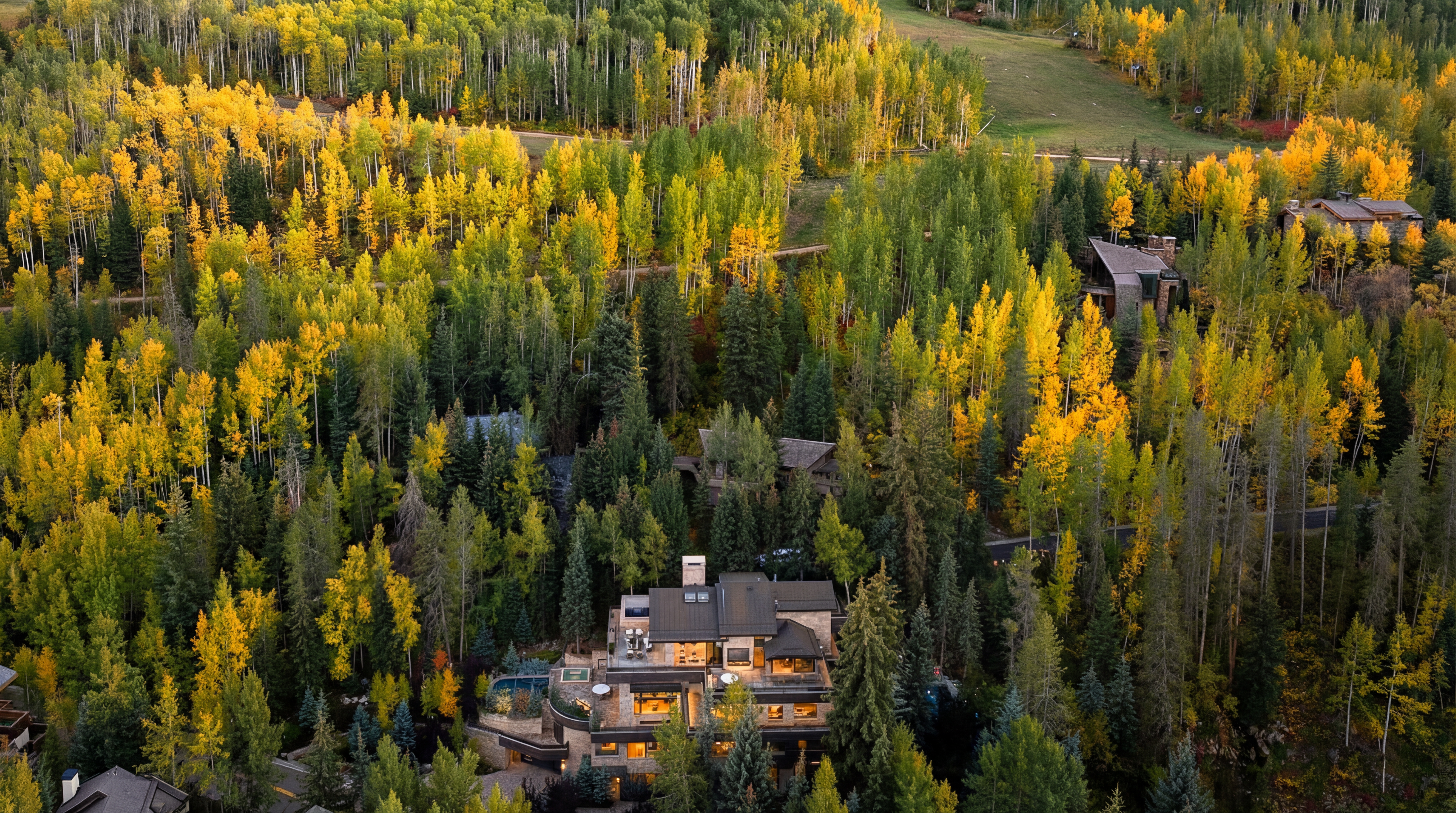 This is an aerial view showcasing a luxury home nestled in a forest with vibrant fall foliage. The house features a modern design with stone accents, a pool, and multiple outdoor living spaces. The image highlights the property's privacy and natural surroundings, emphasizing its high-end appeal.