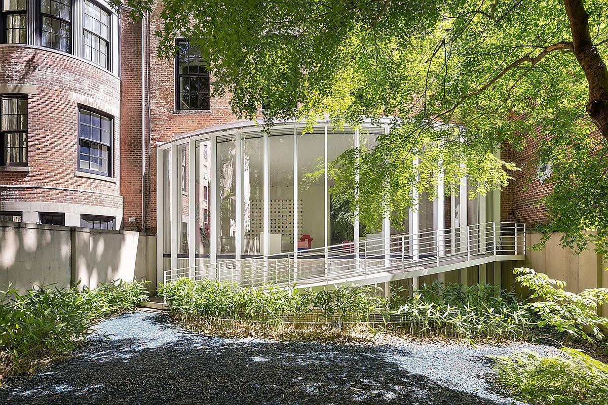 This rear view showcases a striking contrast between a traditional red brick townhouse and a modern, curved glass addition. A metal ramp with a railing leads up to the glass-enclosed space, which is nestled against a lush green tree and a landscaped garden area with dark gravel. The architectural blend of historic masonry and contemporary transparent design creates a unique and sophisticated outdoor aesthetic.
