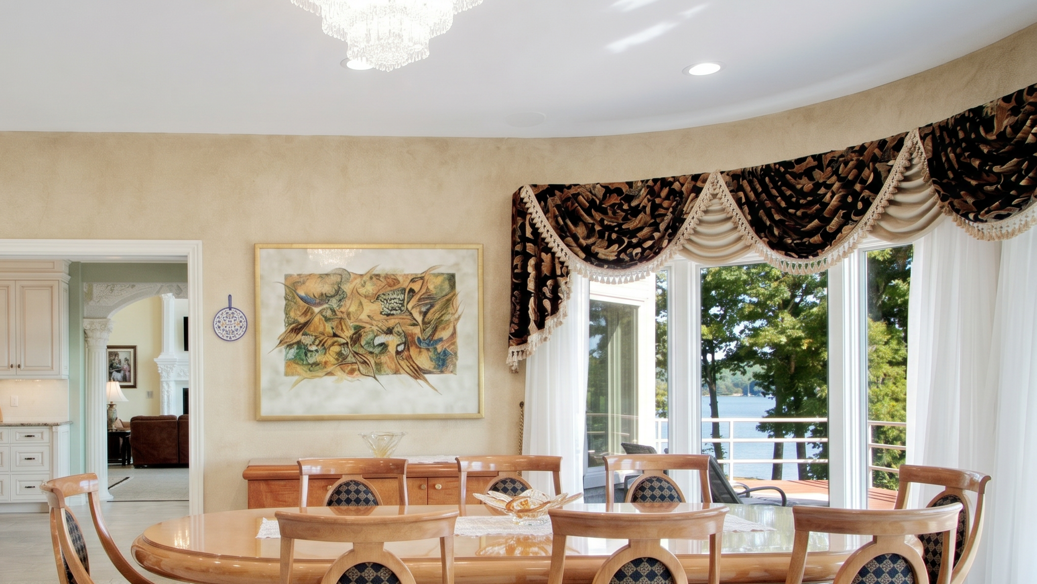 This elegant dining room features a large, polished wood table surrounded by matching chairs with patterned upholstery. The space is beautifully appointed with a crystal chandelier, a large abstract art piece, and dramatic velvet-trimmed window valances that frame a scenic view of the lake. The soft, neutral-toned walls and open transition into a nearby living space create a luxurious and inviting atmosphere for entertaining.