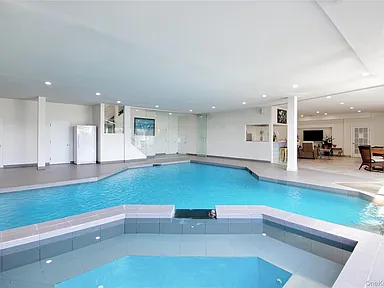 This spacious indoor entertainment area features a large, irregularly shaped indoor swimming pool alongside an adjacent built-in hot tub. The room is designed for leisure with a light, neutral color palette, recessed lighting, and a partially open-concept layout that connects to a living space in the background. It offers a contemporary, multipurpose recreational environment ideal for year-round use.