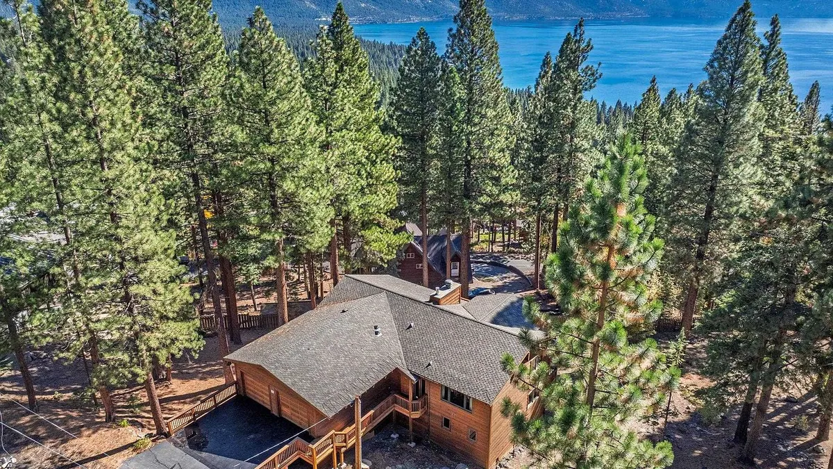 This aerial view showcases a charming mountain home nestled among tall trees, likely pines. The home features a gray shingled roof and wood siding, complemented by a wrap-around deck. The backdrop of a serene lake adds to the property's appeal, highlighting a blend of rustic charm and natural beauty.
