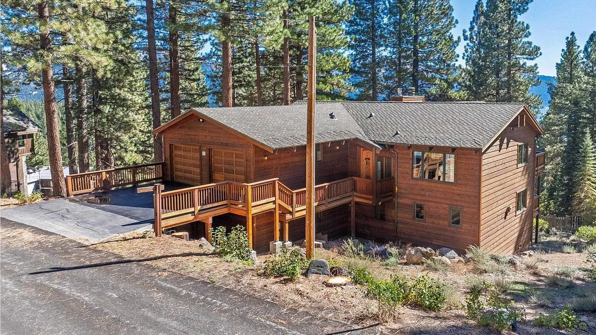This is a front view of a charming wooden house nestled among tall pine trees. A wooden deck with railings leads to the entrance. A garage is attached to the left side of the house with an asphalt driveway leading up to it.