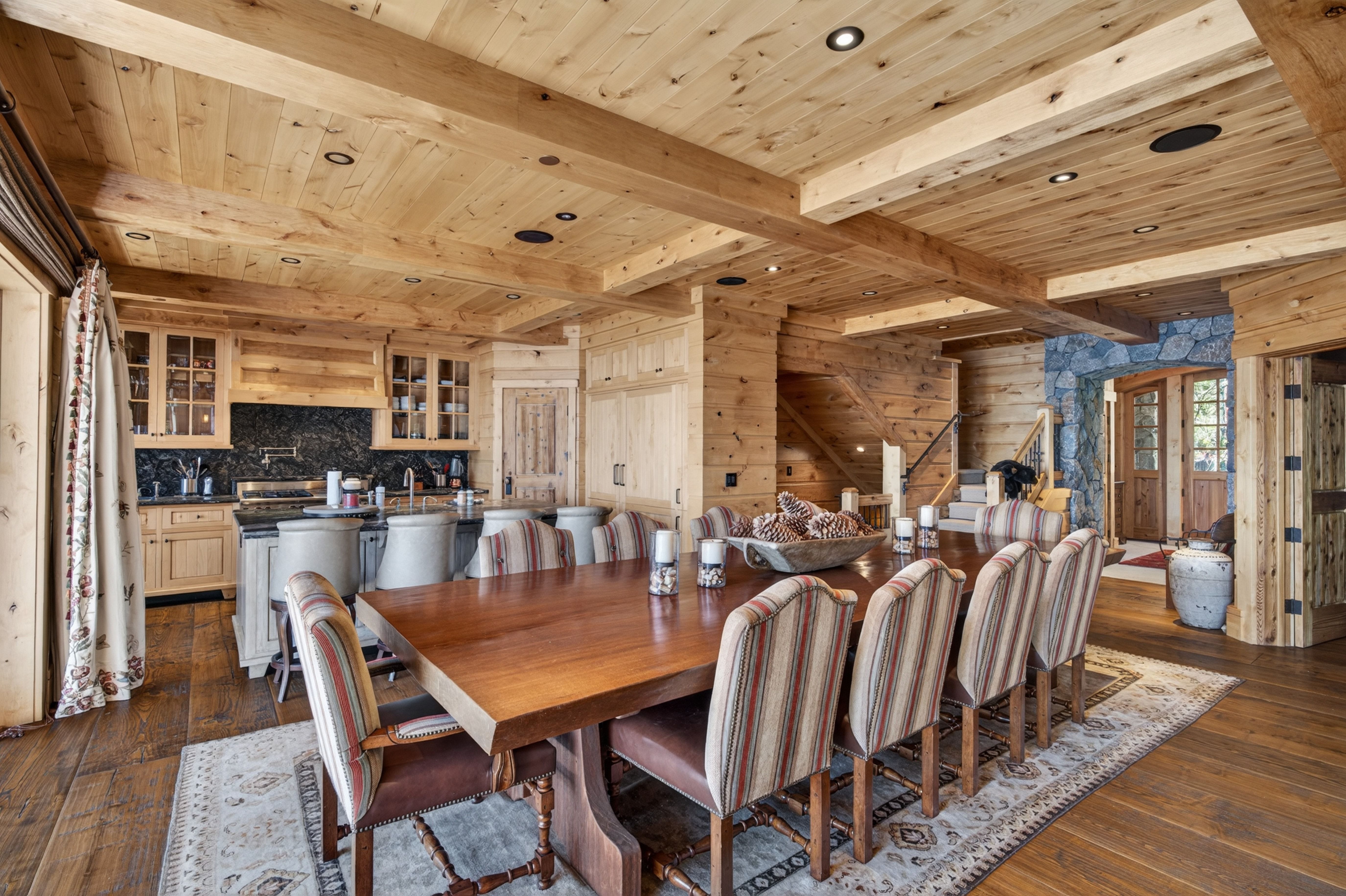 This spacious open-plan dining area features a large, rustic dark wood table surrounded by upholstered chairs with vertical stripes. The room showcases high-end craftsmanship with exposed wooden beam ceilings and matching walls, seamlessly transitioning into an adjacent kitchen with high-quality finishes and dark stone countertops. The area conveys a warm, luxurious cabin atmosphere, bolstered by a decorative rug and natural stone accents near the stairs.