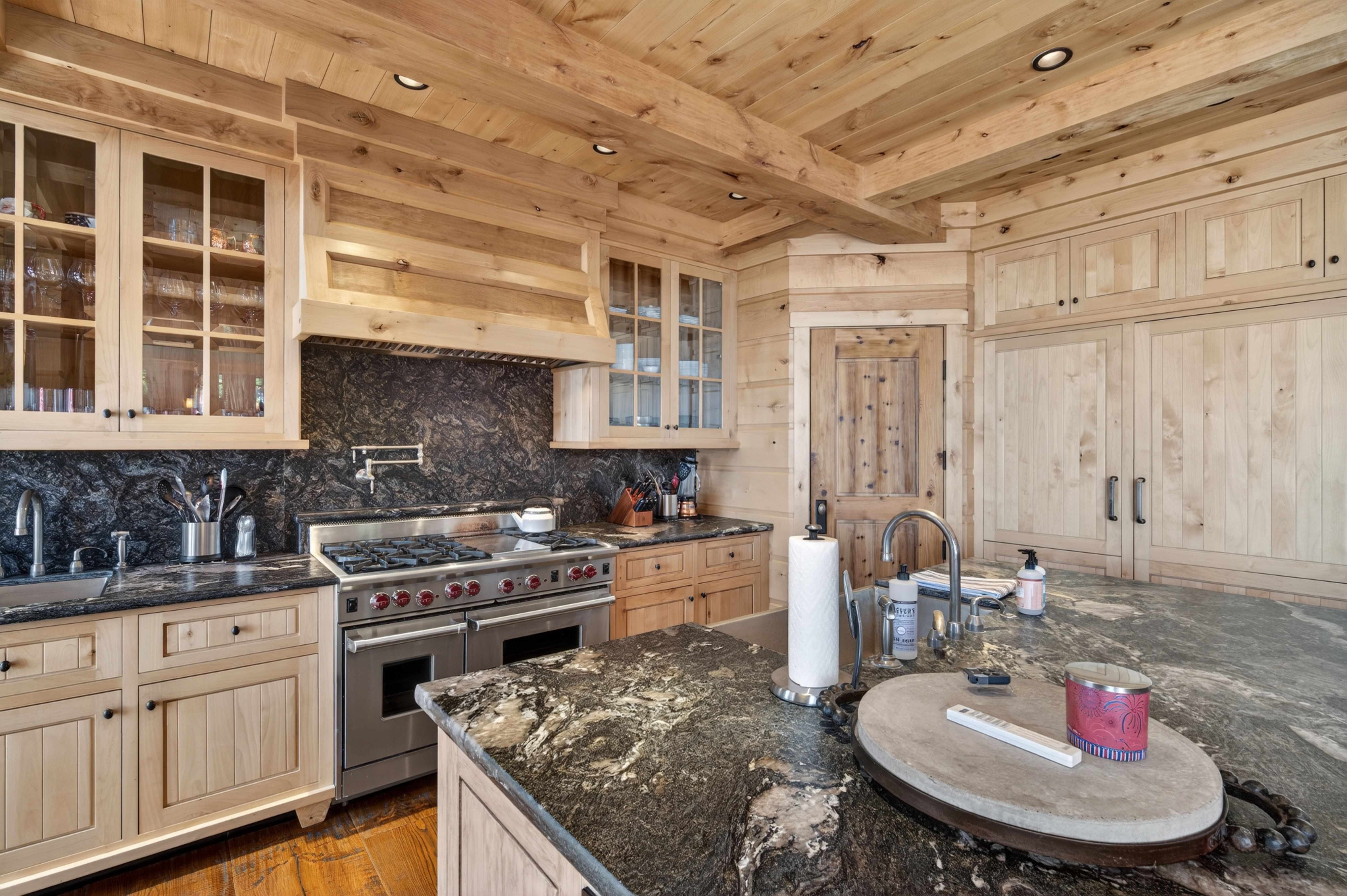 This rustic kitchen showcases high-end craftsmanship with custom light wood cabinetry, exposed wooden ceiling beams, and dramatic black granite countertops. A professional-grade gas range and a large central island with a prep sink highlight the functional layout designed for gourmet cooking. The warm wood tones and rich stone textures create a cozy, luxurious atmosphere characteristic of a high-end mountain home.