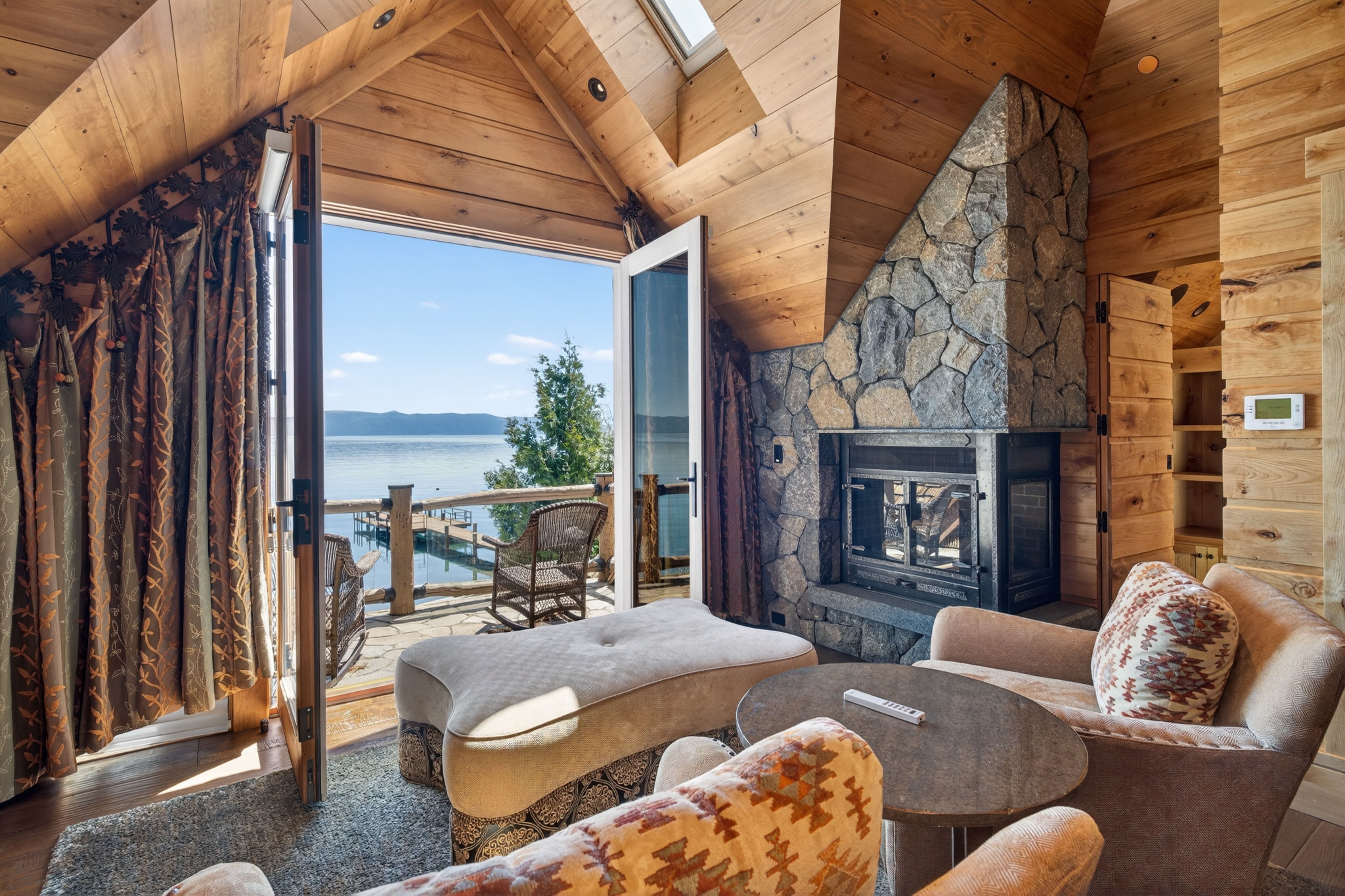 This inviting, rustic living room captures a stunning lakeside view through expansive glass double doors that open onto a private deck with rocking chairs. The space features warm wood-paneled walls and ceilings, complemented by a substantial stone fireplace and comfortable, patterned seating. It offers a cozy, high-end mountain lodge atmosphere perfect for relaxing while enjoying the serene waterfront scenery.