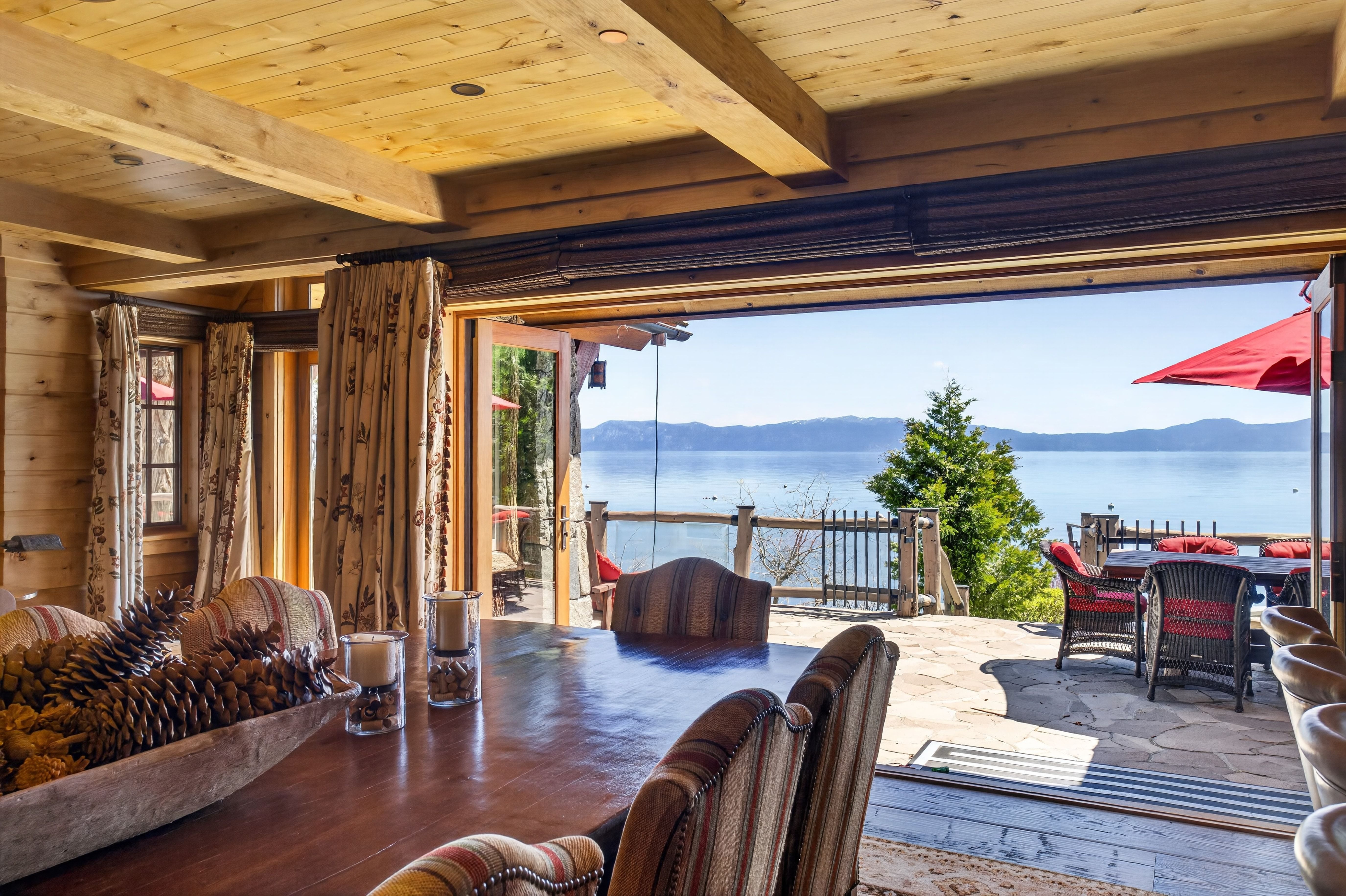 This rustic dining room features a large, rich wood table set against a backdrop of a stunning, panoramic view of a calm lake and distant mountains. Large open folding glass doors blur the lines between indoor and outdoor living, leading onto a stone patio equipped with comfortable seating. The heavy wood-beamed ceiling and warm wood walls create a cozy, lodge-like aesthetic that emphasizes the luxurious lakeside setting.