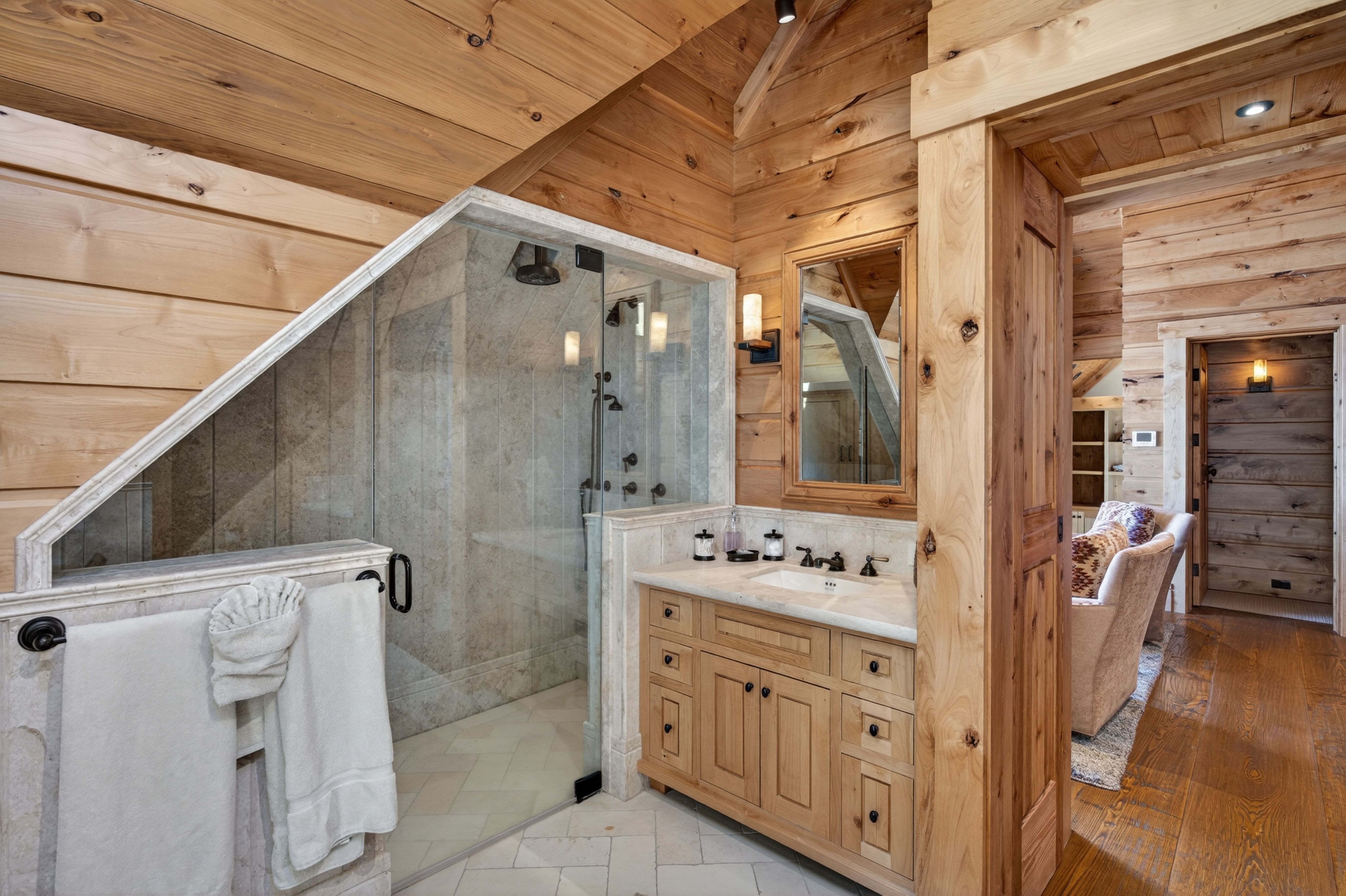 This rustic primary bathroom features warm wood-paneled walls and ceilings, creating a cozy cabin aesthetic. The space includes a glass-enclosed, tiled walk-in shower and a custom wooden vanity with a stone countertop. Elegant details, such as antique-style hardware and soft interior lighting, complement the natural textures of the room.