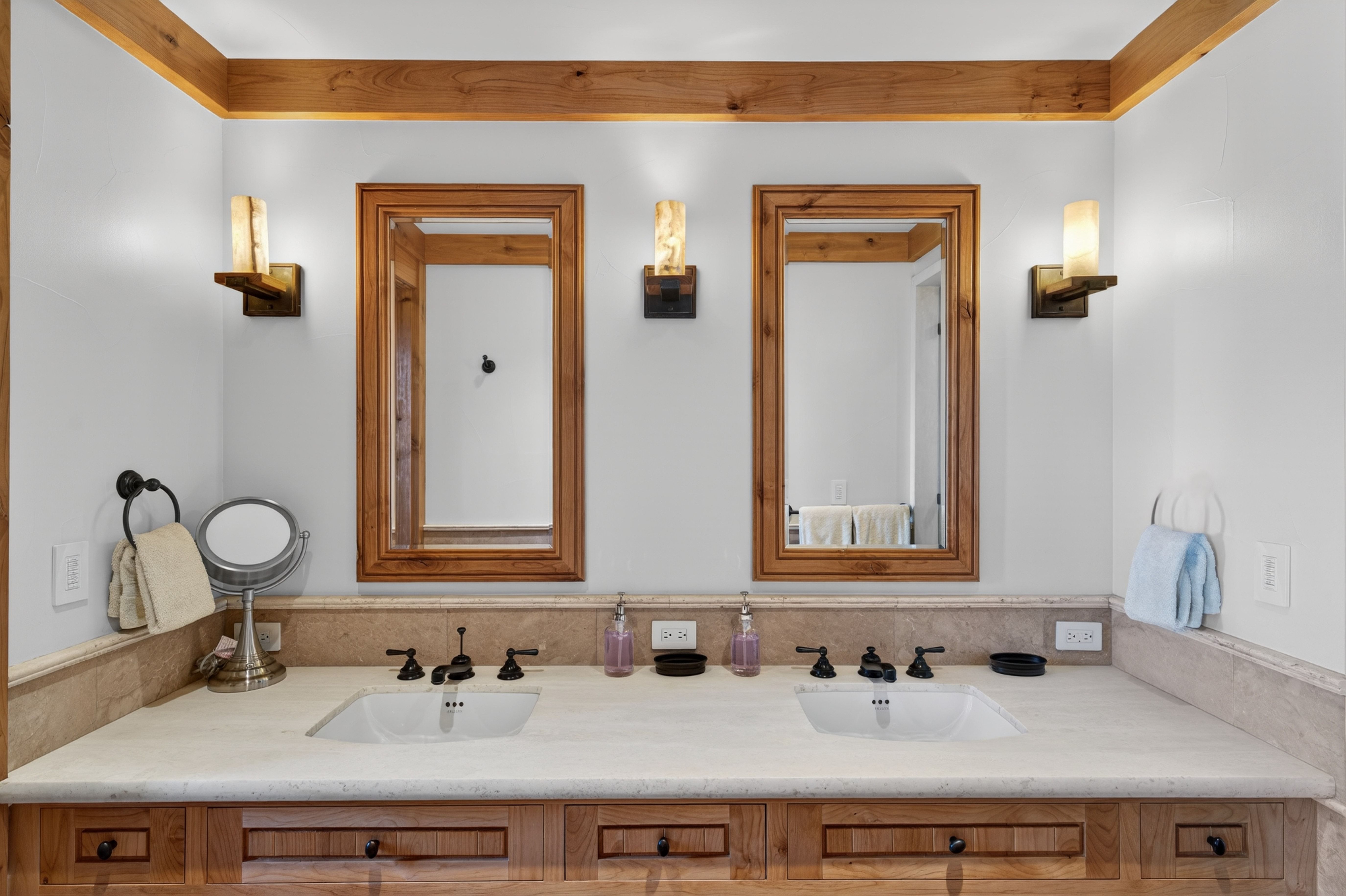 This primary bathroom features a rustic, dual-sink vanity with warm wood cabinetry and a light stone countertop. Twin framed mirrors are accented by elegant sconce lighting, while the neutral color palette with stone backsplash creates a clean and sophisticated atmosphere. The overall design feels upscale and cohesive, perfect for a high-end mountain or craftsman-style home.