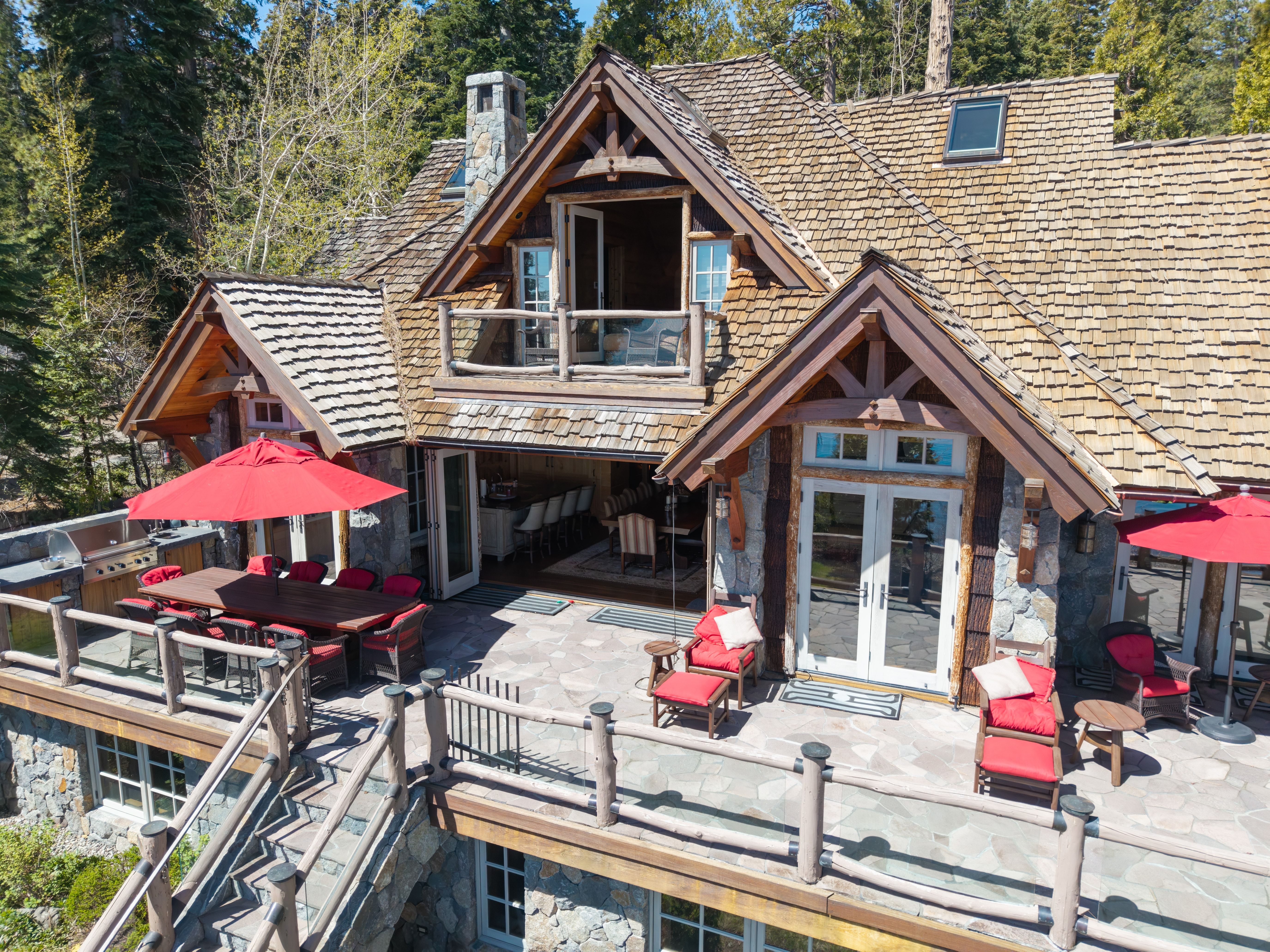 This elevated rear view of a rustic mountain home highlights a spacious stone patio perfect for outdoor entertaining. The home features classic timber framing, shingle siding, and a large stone chimney, paired with expansive glass doors that create a seamless transition between indoor and outdoor living. The deck is well-appointed with red-cushioned seating and dining areas, all framed by the lush forest backdrop.