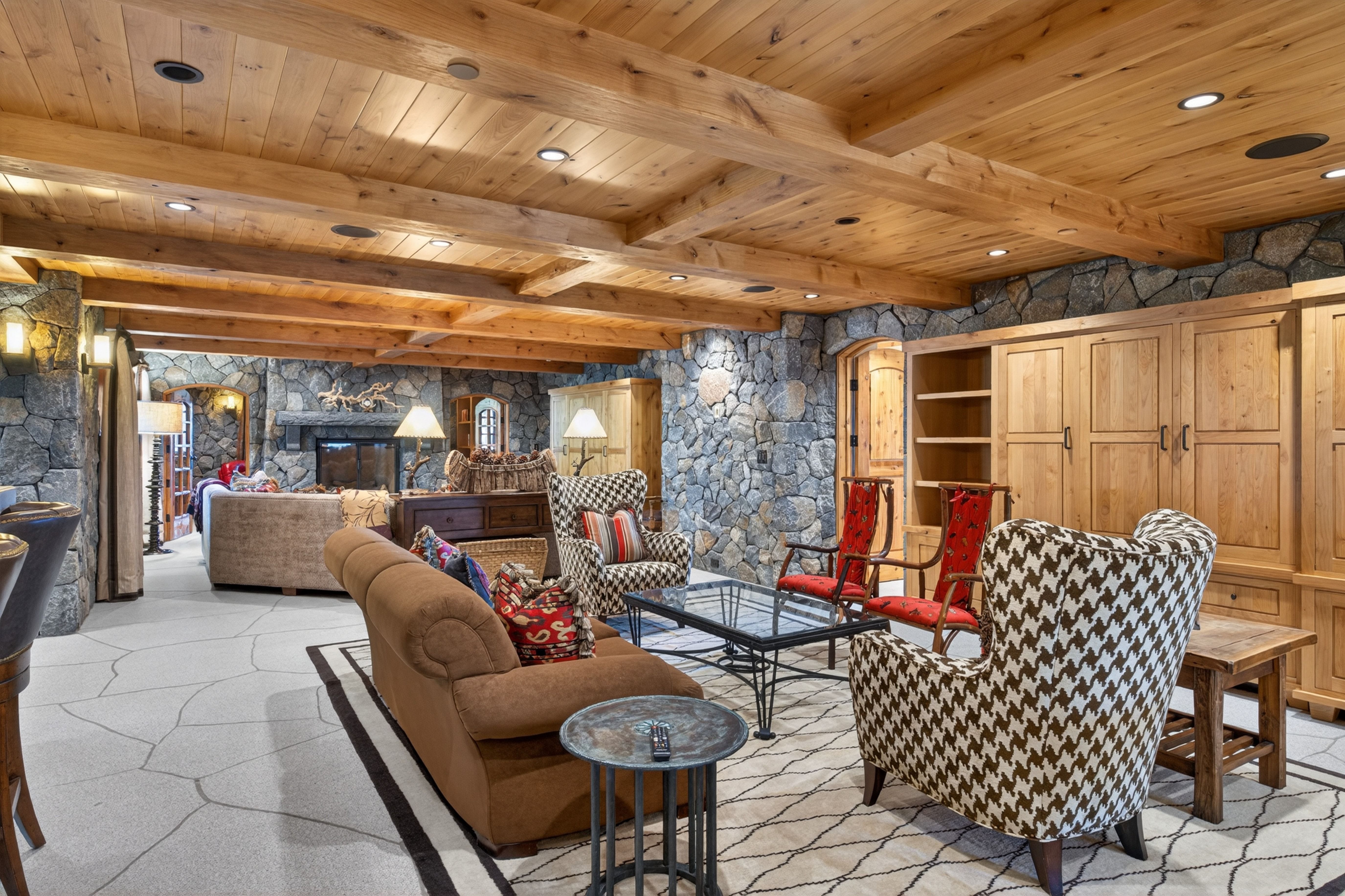 This spacious living area features a warm, rustic aesthetic with exposed timber beamed ceilings and dramatic natural stone walls. The room is furnished with a mix of comfortable sofas and wingback chairs, centered around a glass-topped coffee table on a patterned area rug. Large built-in wood cabinetry and elegant decor create an inviting, cabin-inspired atmosphere perfect for relaxation.