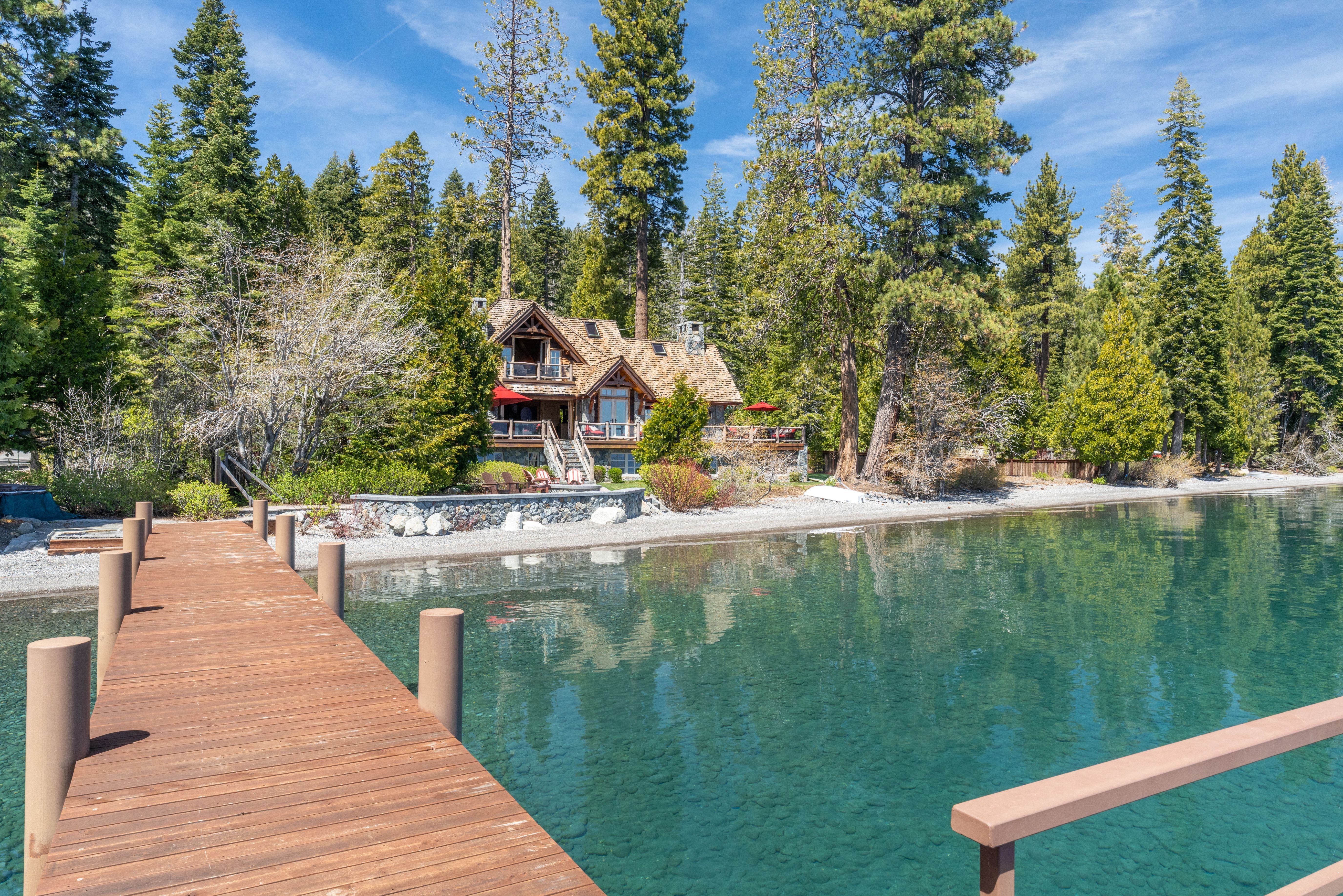 This stunning lakeside property features a rustic, multi-story timber-frame home nestled amongst a forest of towering pine trees. A private wooden pier leads out into the crystal-clear water, while the home impresses with large decks, elegant wood and stone architecture, and stone landscaping that integrates perfectly with the shoreline. It presents an idyllic, high-end waterfront retreat characterized by serene natural beauty.