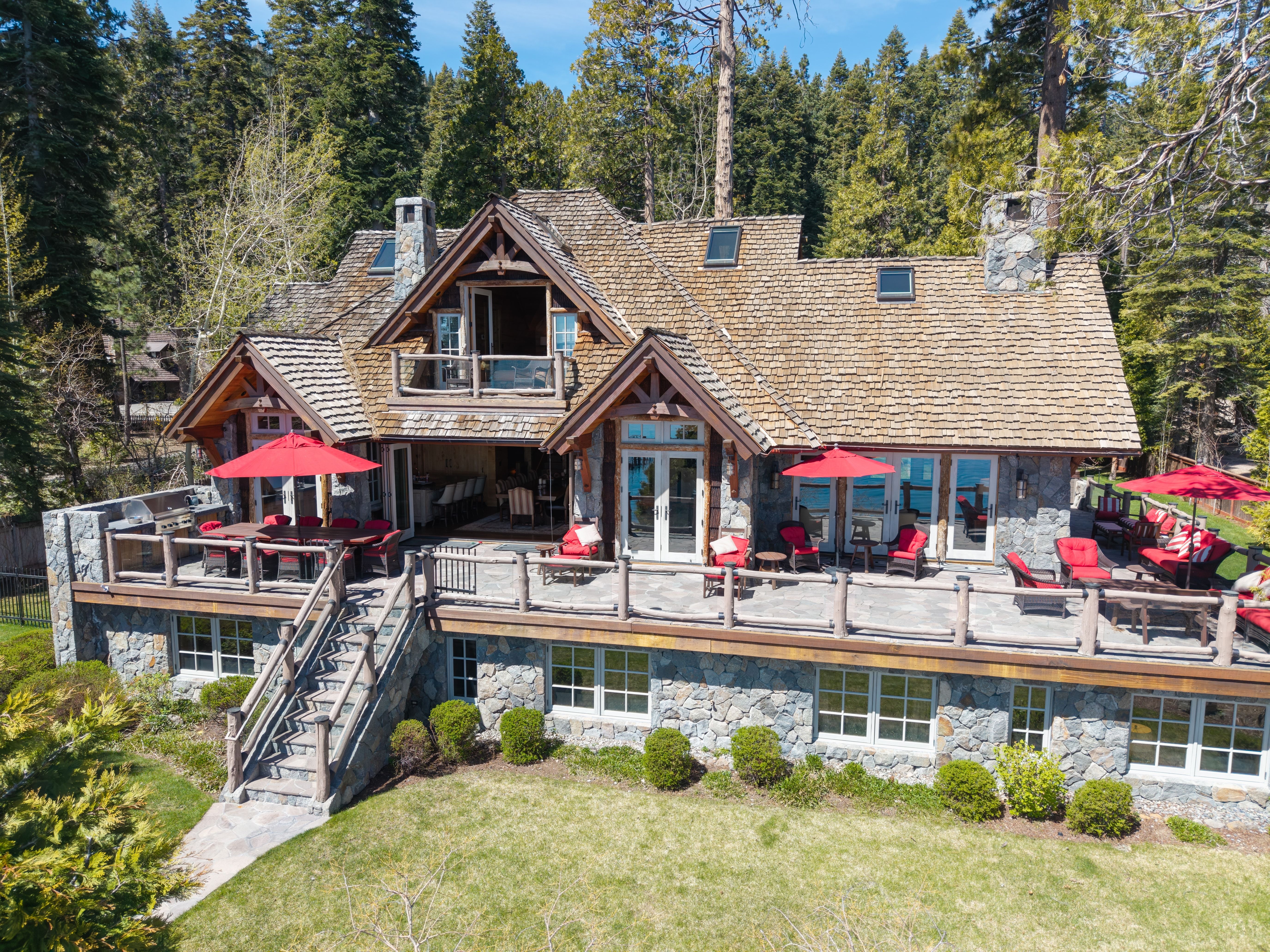 This stunning mountain-style home showcases craftsman architecture with a rugged stone facade and wood shingle roofing. The expansive multi-level stone patio and deck feature comfortable outdoor seating with red accents, perfect for taking in the scenic wooded views. The home's design blends seamlessly into the natural, lush forest landscape, creating a luxurious and serene retreat.