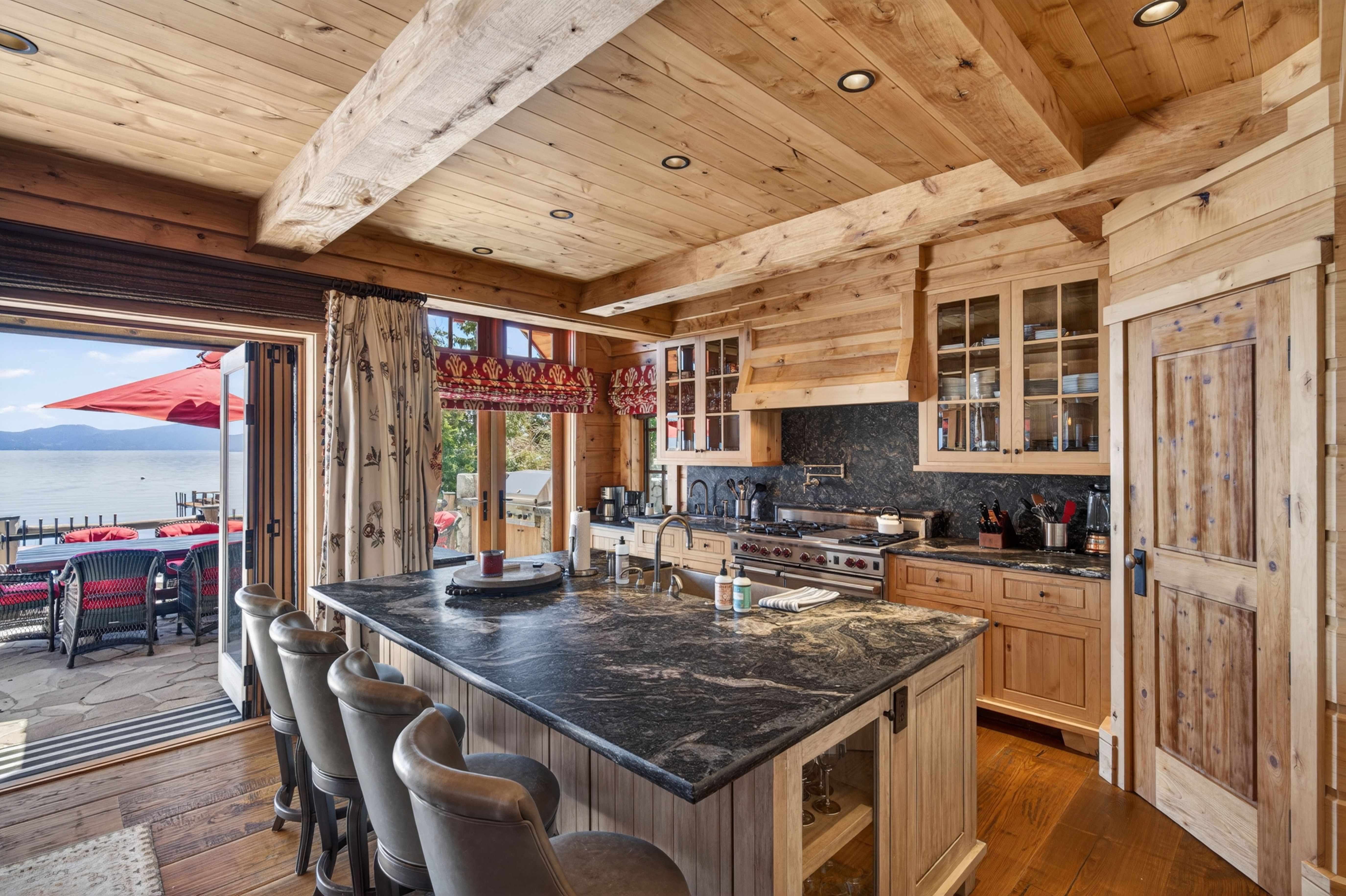 This rustic, high-end kitchen features natural wood paneling, exposed beams, and a large kitchen island topped with dark, veined stone countertops. The space seamlessly integrates indoor and outdoor living with wide-opening patio doors that provide direct access to a deck overlooking a scenic lake. With its warm cabinetry, professional-grade range, and abundant natural light, the kitchen offers both luxury appeal and a cozy, cabin-inspired aesthetic.
