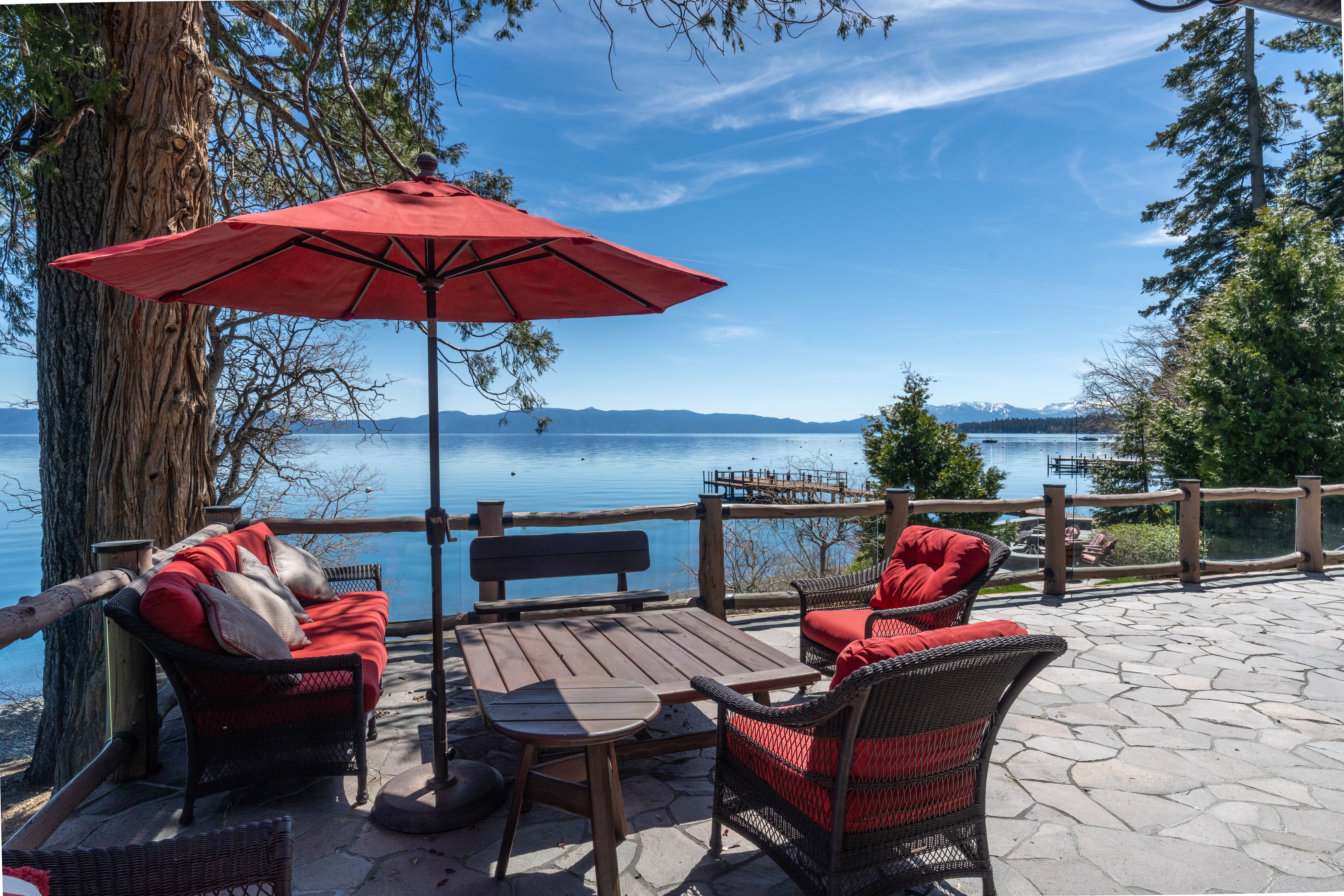 This serene waterfront patio provides an exceptional outdoor living space, featuring high-quality wicker furniture with vibrant red cushions and a large umbrella for shade. The stone-paved deck overlooks a tranquil lake and mountains, offering stunning, unobstructed natural views. It is an ideal setting for relaxation or entertaining in a scenic, peaceful environment.