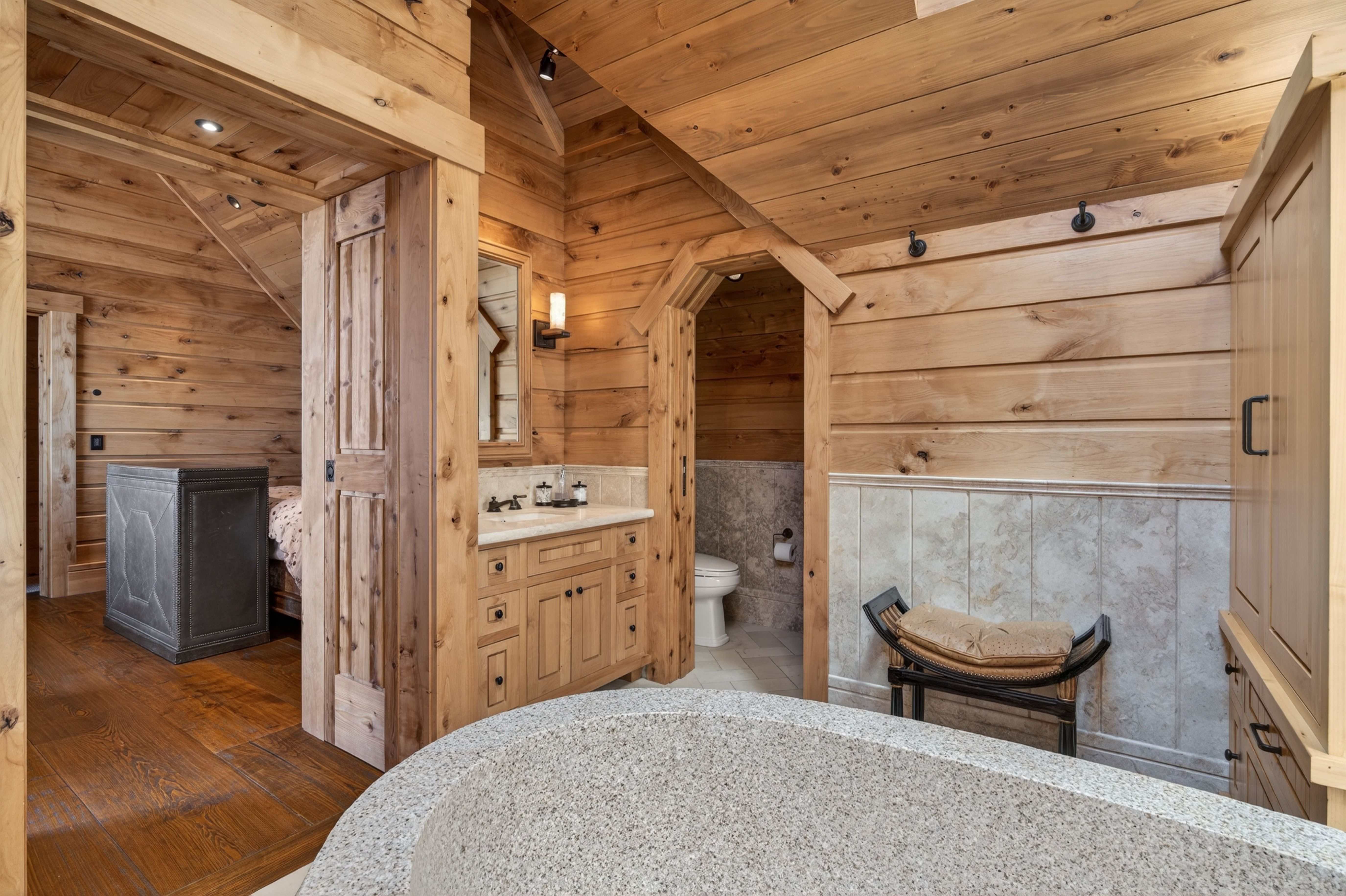 This rustic primary bathroom features warm wood-paneled walls and a ceiling, providing a cozy, cabin-like aesthetic. It is equipped with a custom stone bathtub, a wooden vanity with a stone countertop, and a separate private area for the toilet. A small bench with a neutral cushion adds seating, while the overall design elegantly blends natural stone textures with rich timber elements.