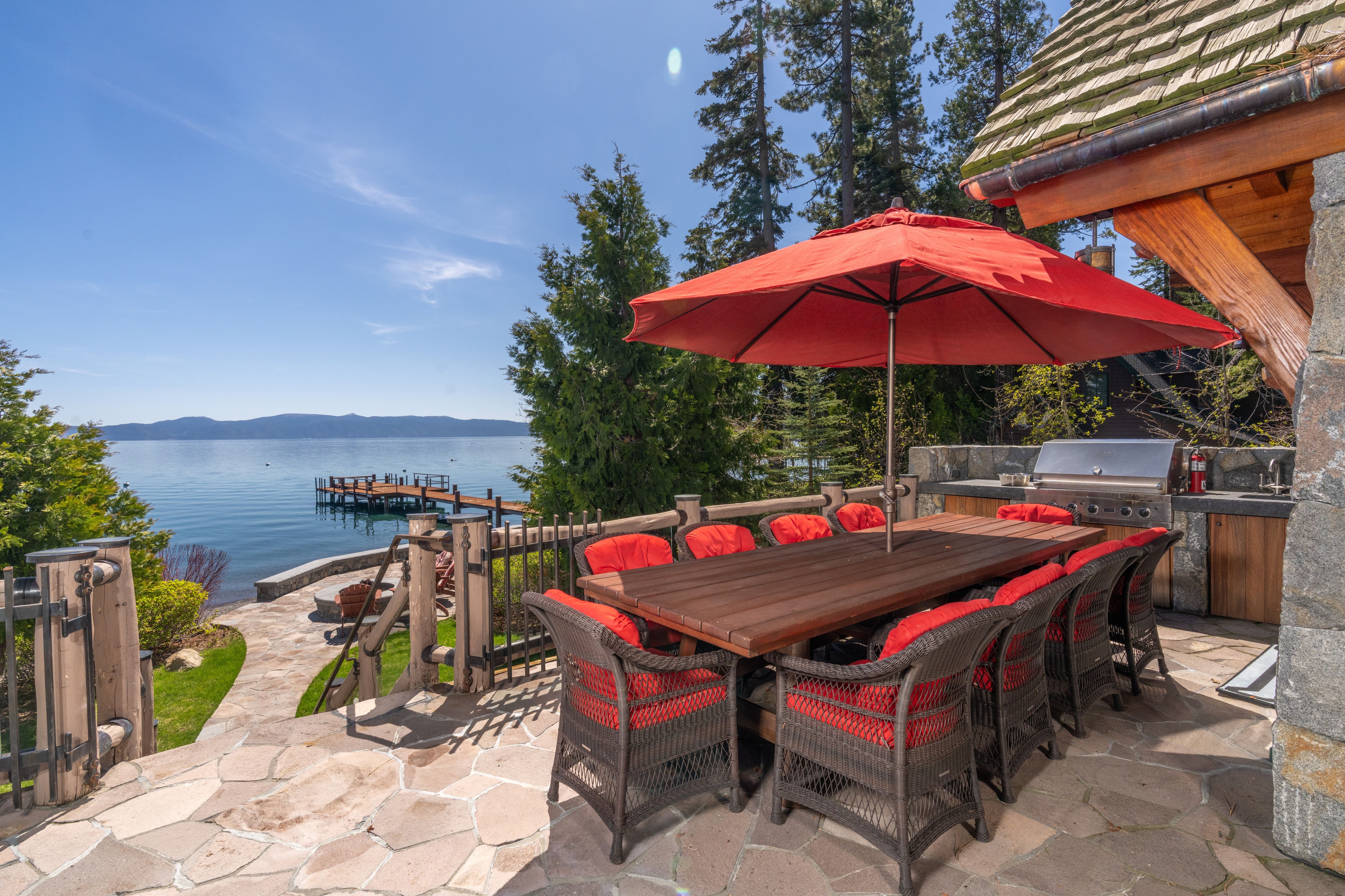 This scenic stone patio features an outdoor dining set with a large red umbrella, perfect for lakefront entertaining. The space includes a built-in outdoor grill area and offers stunning, direct views of the lake and a private dock. It provides a luxurious and relaxing atmosphere, ideal for enjoying lakeside living.