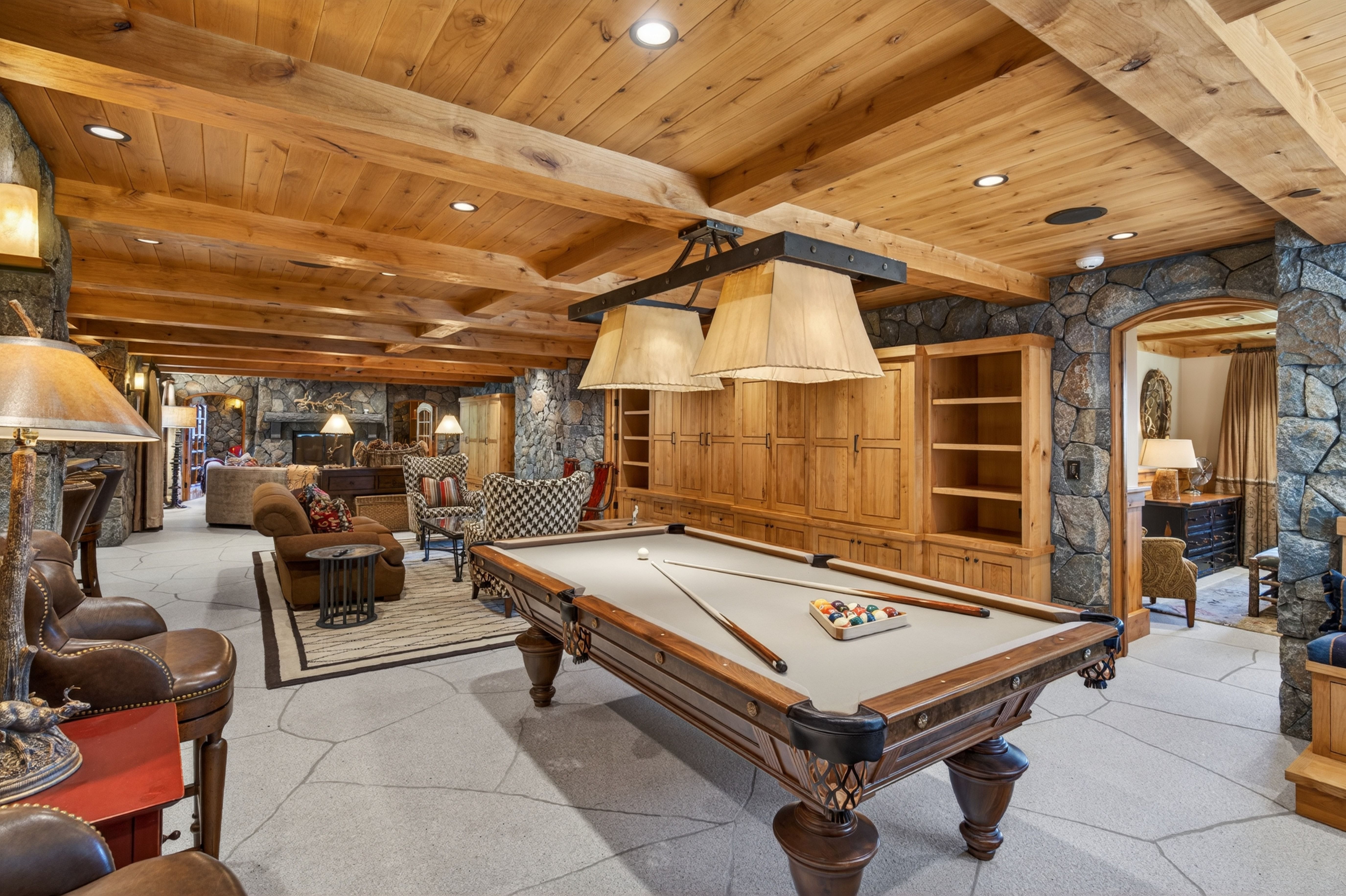 This spacious and rustic finished basement features beautiful exposed wood beam ceilings, stone accents, and custom built-in cabinetry. The room is well-appointed with a billiards table as a central focal point, leading into a comfortable lounge area with plush seating, making it an ideal space for entertaining. The open-concept design combined with natural textures and a cohesive color palette creates a cozy and inviting atmosphere.