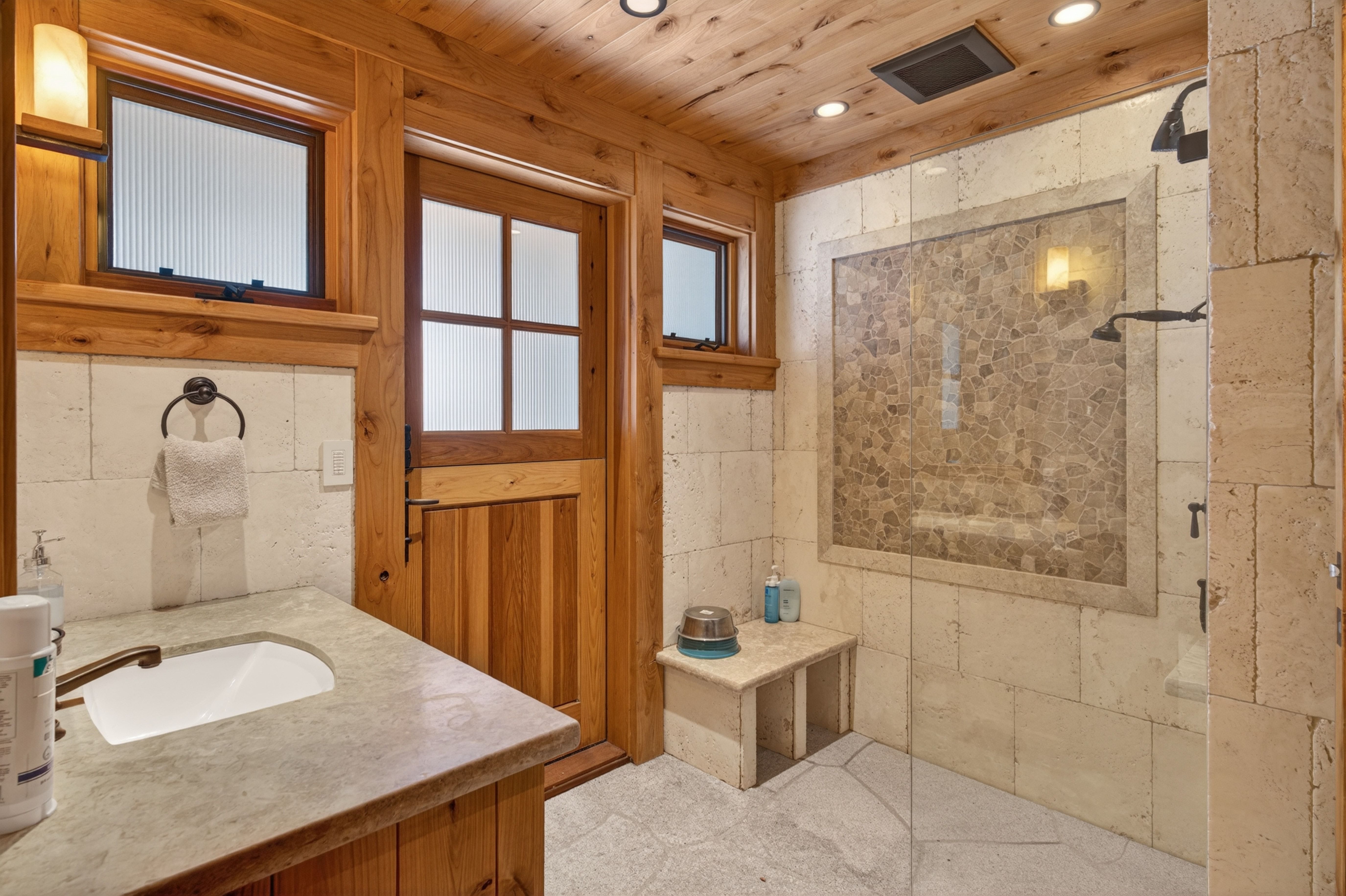 This rustic, lodge-style bathroom features warm wood-paneled ceilings and door frames combined with natural stone textures throughout. Key elements include a walk-in shower with stone tile inlay, a built-in stone bench, and an integrated stone countertop with a sleek white vanity sink. The use of natural materials creates a cozy, spa-like atmosphere consistent with mountain or cabin aesthetics.