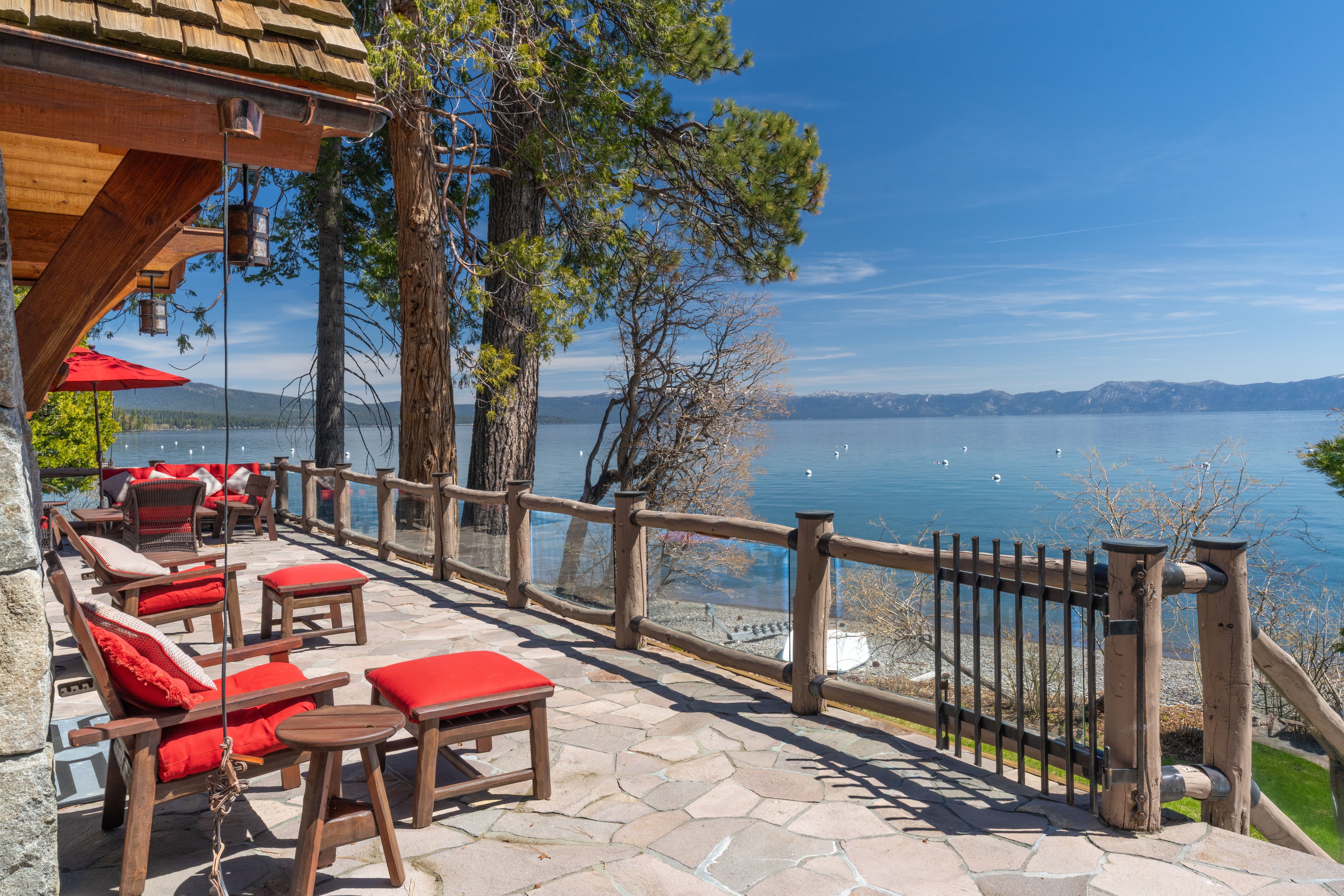This spacious, stone-paved lakeside deck features comfortable rustic seating with bright red cushions and a matching umbrella, perfect for outdoor relaxation. Enclosed by a natural wood and glass railing that preserves the unobstructed scenic view, the deck offers a stunning perspective of the water, distant mountains, and surrounding pine trees. It provides a luxurious, tranquil atmosphere ideal for enjoying the picturesque shoreline.