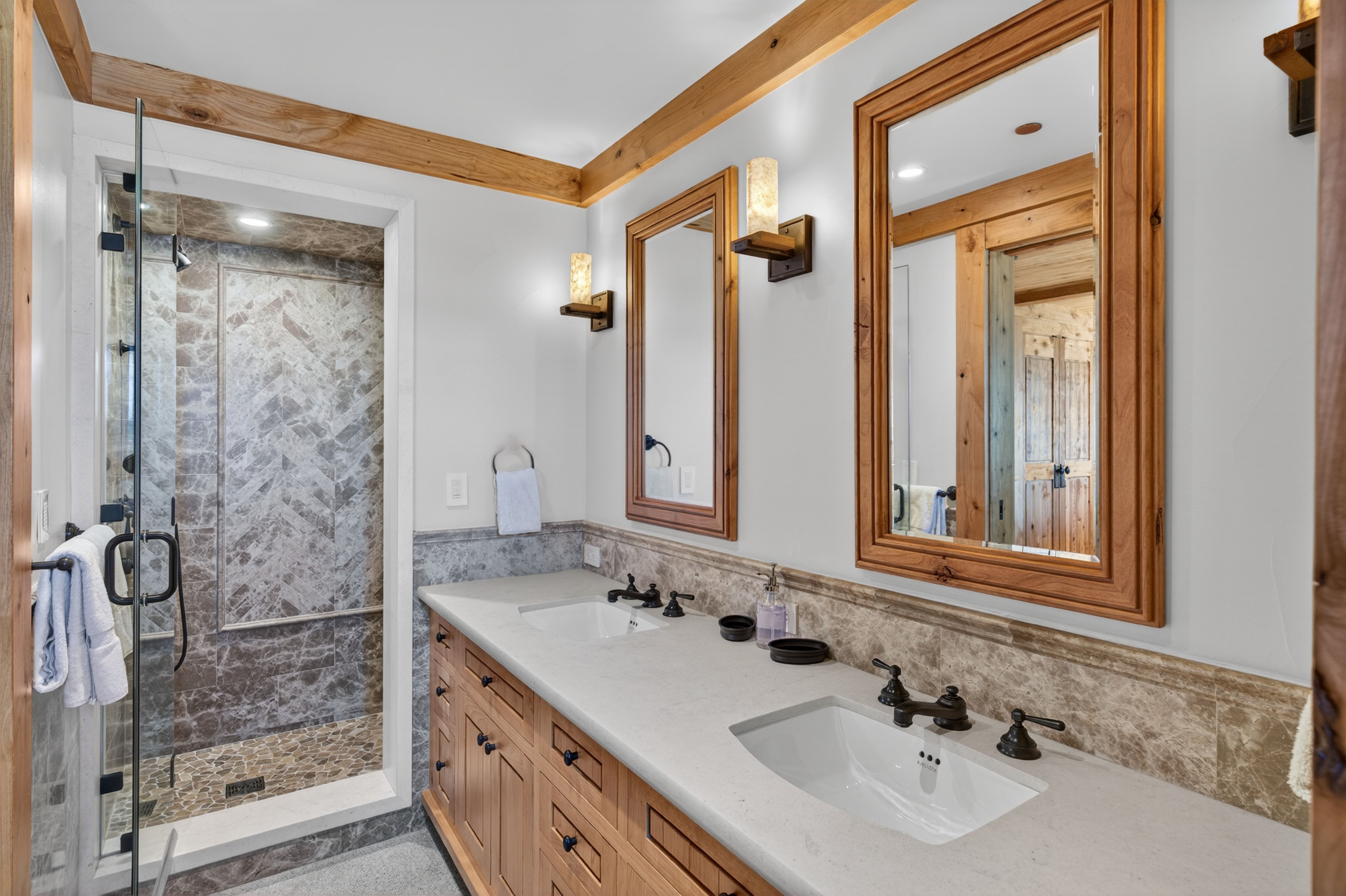 This primary bathroom features a rustic wood-framed vanity with a double sink, topped by light quartz countertops and elegant oil-rubbed bronze fixtures. A glass-enclosed walk-in shower showcases intricate marble tile work in a herringbone pattern and a natural stone floor. The space is tied together with warm wood trim accents, natural-toned stone backsplash, and matching wall sconces, creating a luxurious mountain lodge aesthetic.