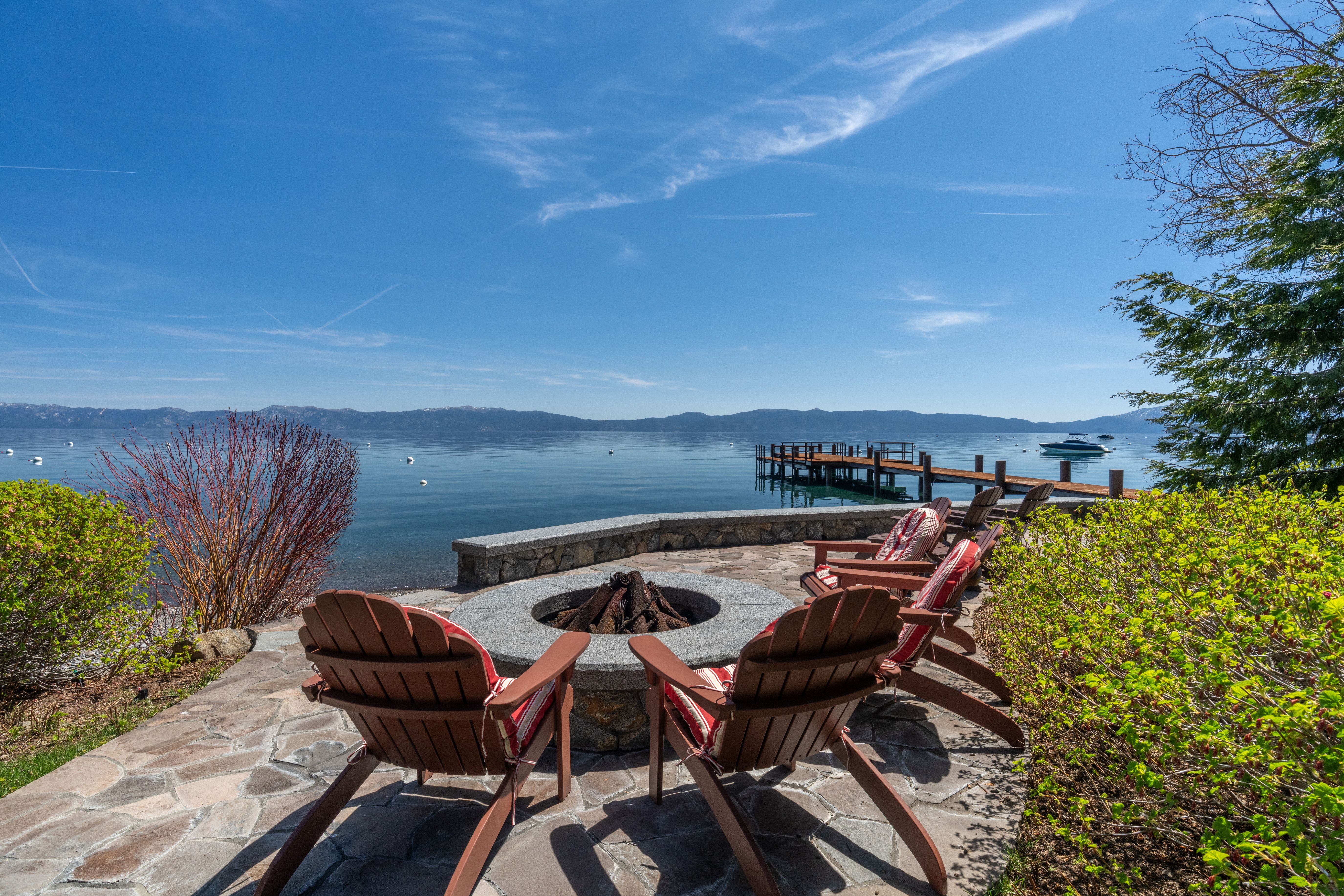This stunning waterfront patio features a flagstone surface arranged around a stone fire pit, complete with Adirondack-style chairs featuring striped cushions. The area offers panoramic, unobstructed views of the lake and mountains, highlighted by a private wooden dock extending into the water. Its serene, upscale setting provides an ideal space for outdoor relaxation and entertaining.