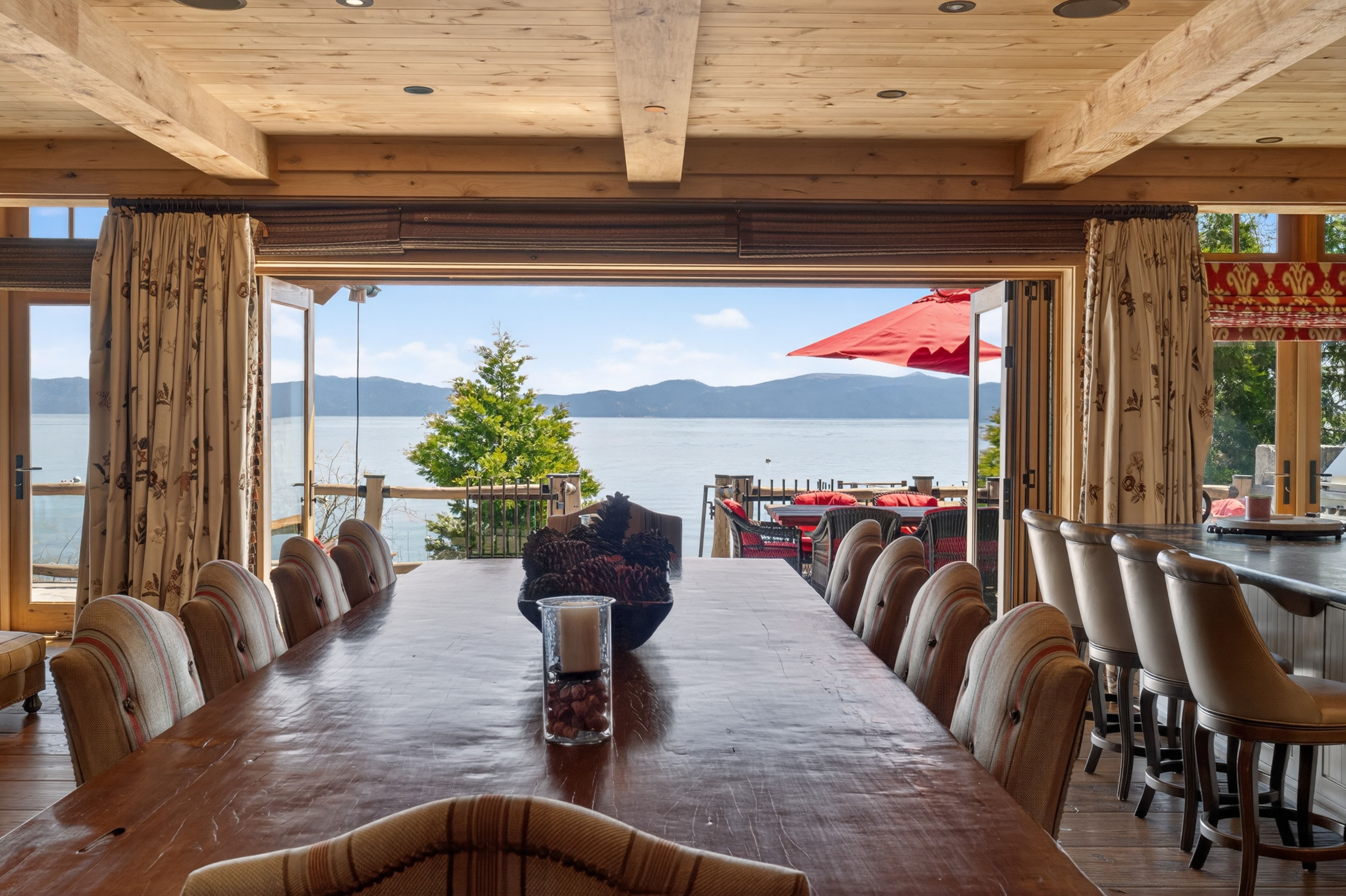 This spacious dining area features a large, rustic wooden table and comfortable, patterned upholstered chairs, perfect for hosting family gatherings. The room seamlessly flows into a massive outdoor deck through wide-opening doors, revealing breathtaking panoramic views of a tranquil lake and distant mountains. Complementing the natural beauty outside, the interior boasts warm wood-beamed ceilings and a cozy, elevated cabin aesthetic.