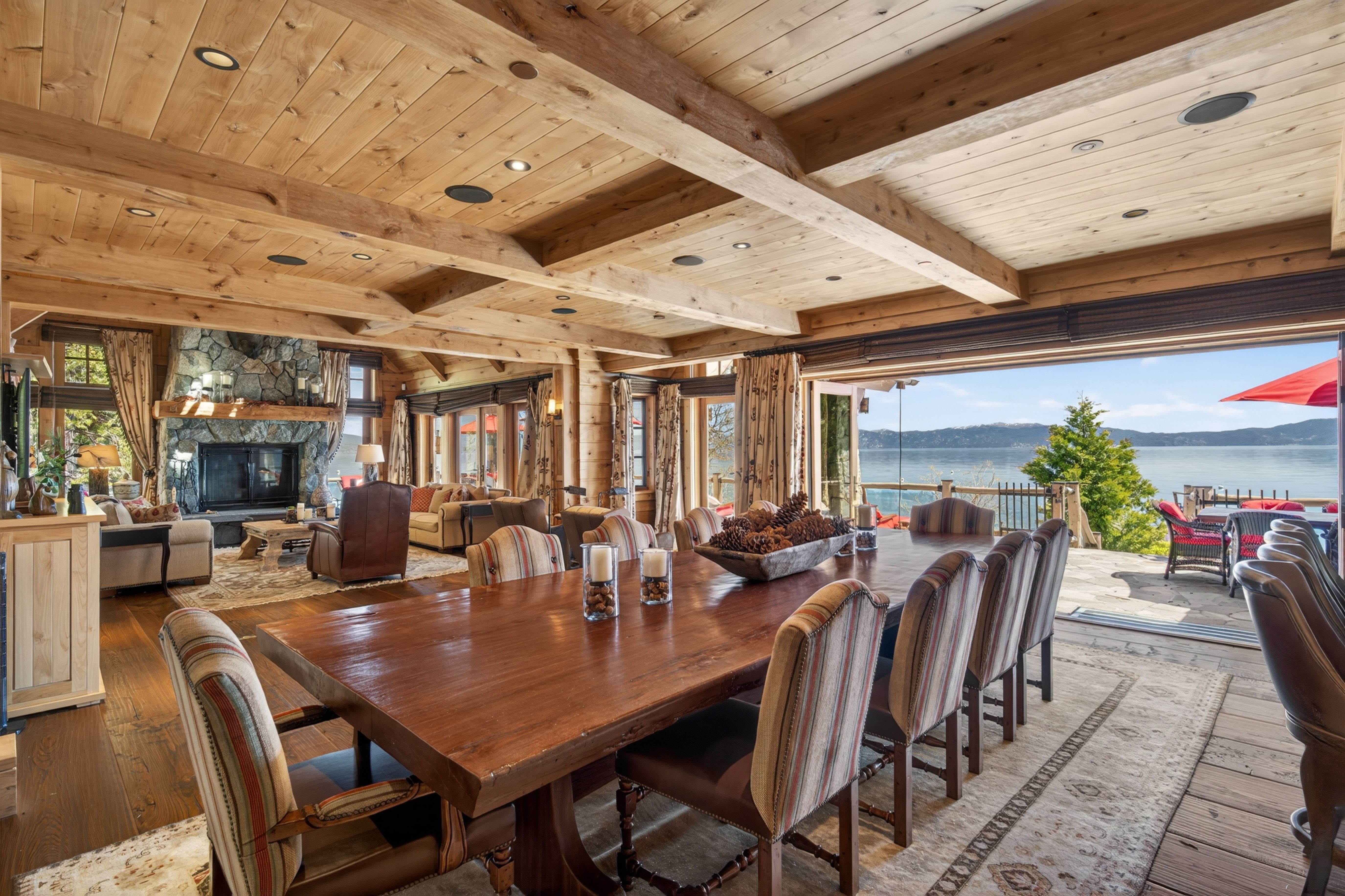 This elegant, lodge-style dining area features a massive, dark wood dining table surrounded by high-backed, striped upholstered chairs, perfect for large gatherings. The open-concept space flows seamlessly into a cozy living area with a stone fireplace and leads to a sprawling deck through floor-to-ceiling glass doors, offering panoramic lake views. Vaulted wood-beam ceilings and refined rustic decor create a warm, luxurious atmosphere characteristic of premium mountain estates.