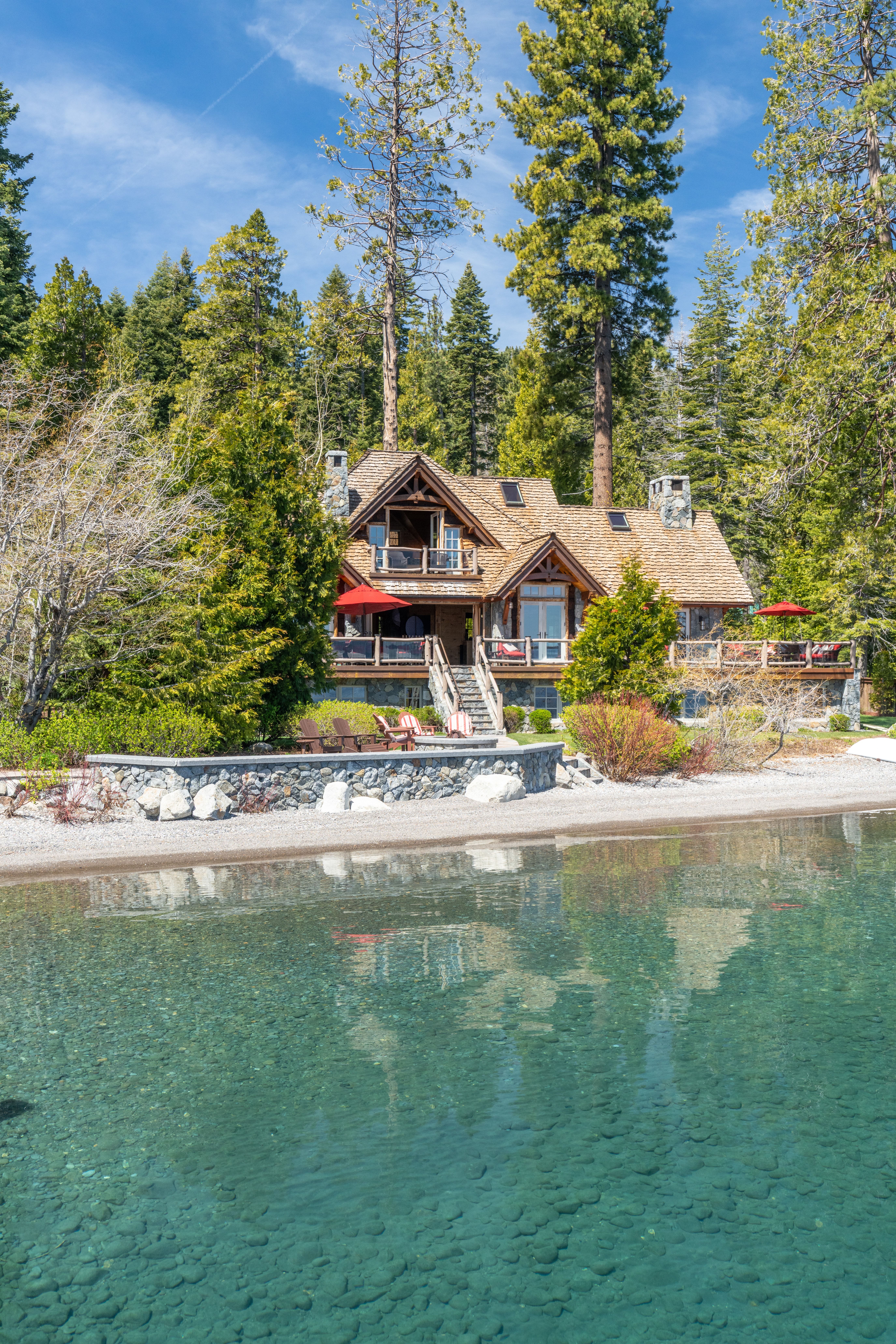 This scenic lakefront property showcases a charming, rustic-style cabin constructed with natural stone and wood textures. The exterior features a prominent deck with multiple levels, expansive seating areas, and red umbrellas for shade, overlooking a crystal-clear pebble-bottom shoreline. Surrounded by dense pine trees, the home offers a serene and secluded atmosphere perfect for waterfront living.