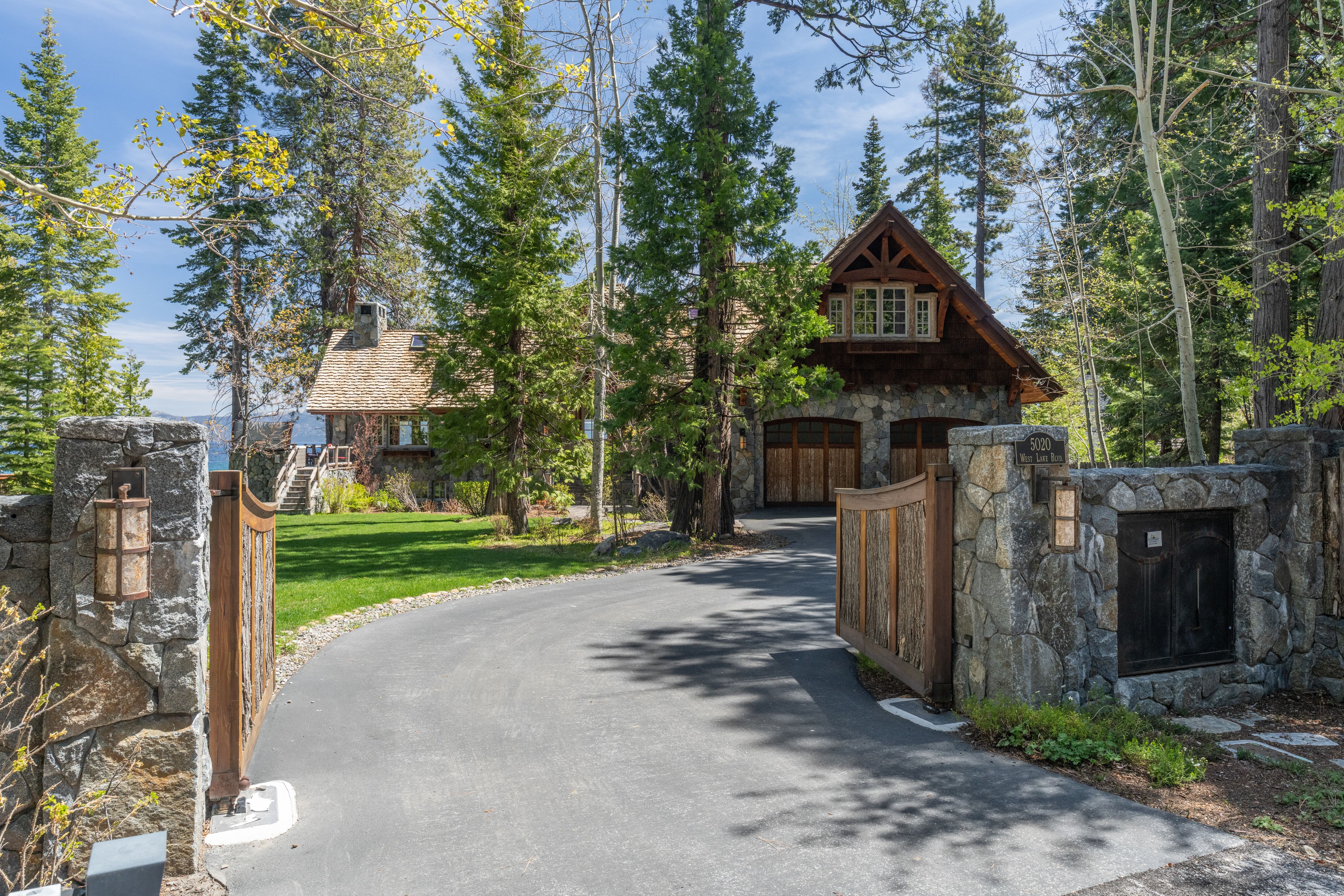 This image showcases a stunning luxury estate entryway, framed by stone pillars and custom wooden gates. The home features classic timber-frame and stone mountain architecture, set against a backdrop of lush pine trees and a well-manicured lawn with a paved driveway leading to a multi-car garage. It provides an inviting and prestigious impression, characteristic of high-end mountain retreats.
