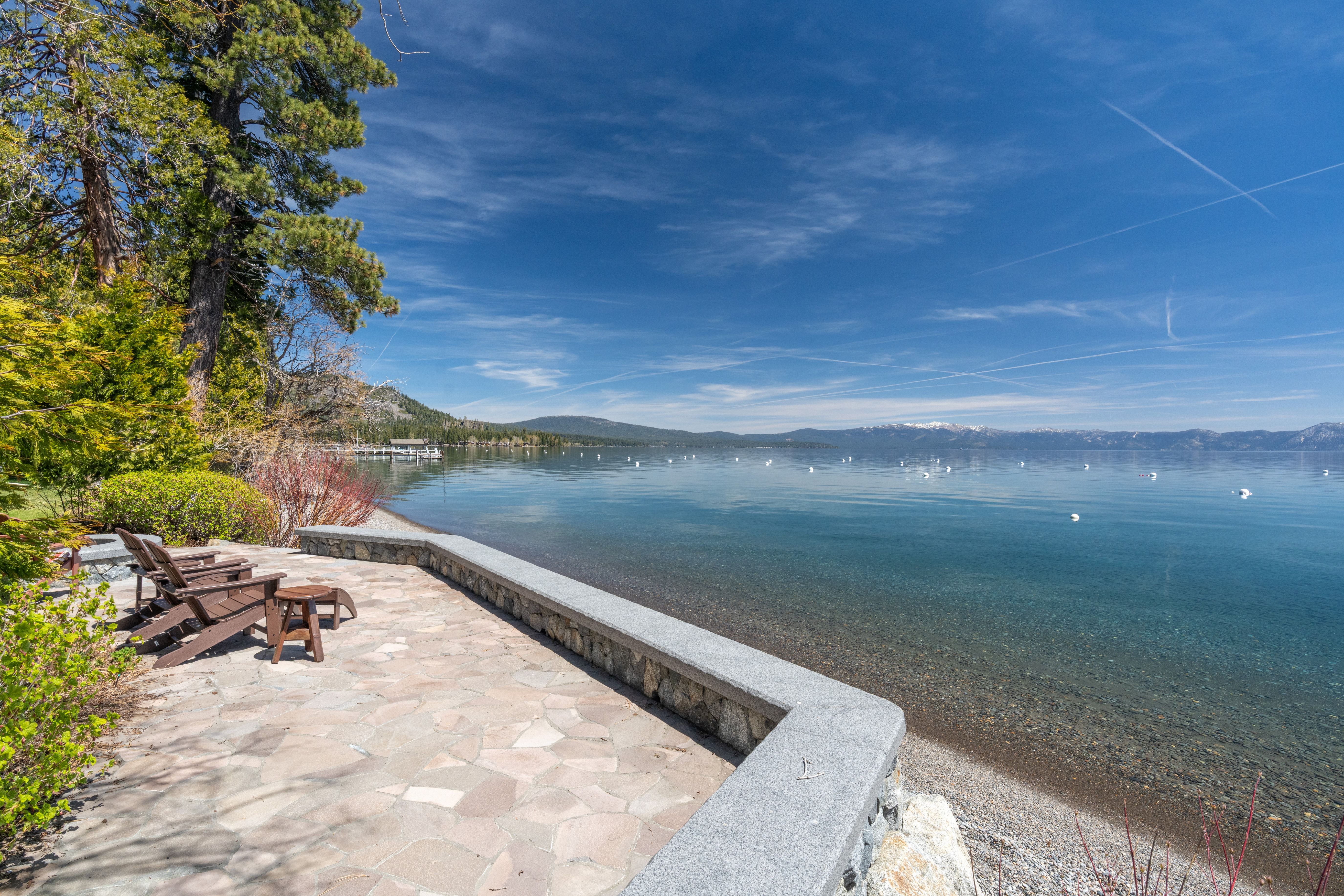 This stunning waterfront patio features natural stone paving and a sleek stone retaining wall that overlooks a crystal-clear lake. Adirondack chairs with a cocktail table are perfectly positioned to enjoy the serene, scenic view of the lake and distant mountains. The space offers an inviting and tranquil outdoor living area, ideal for relaxing or entertaining right at the shoreline.