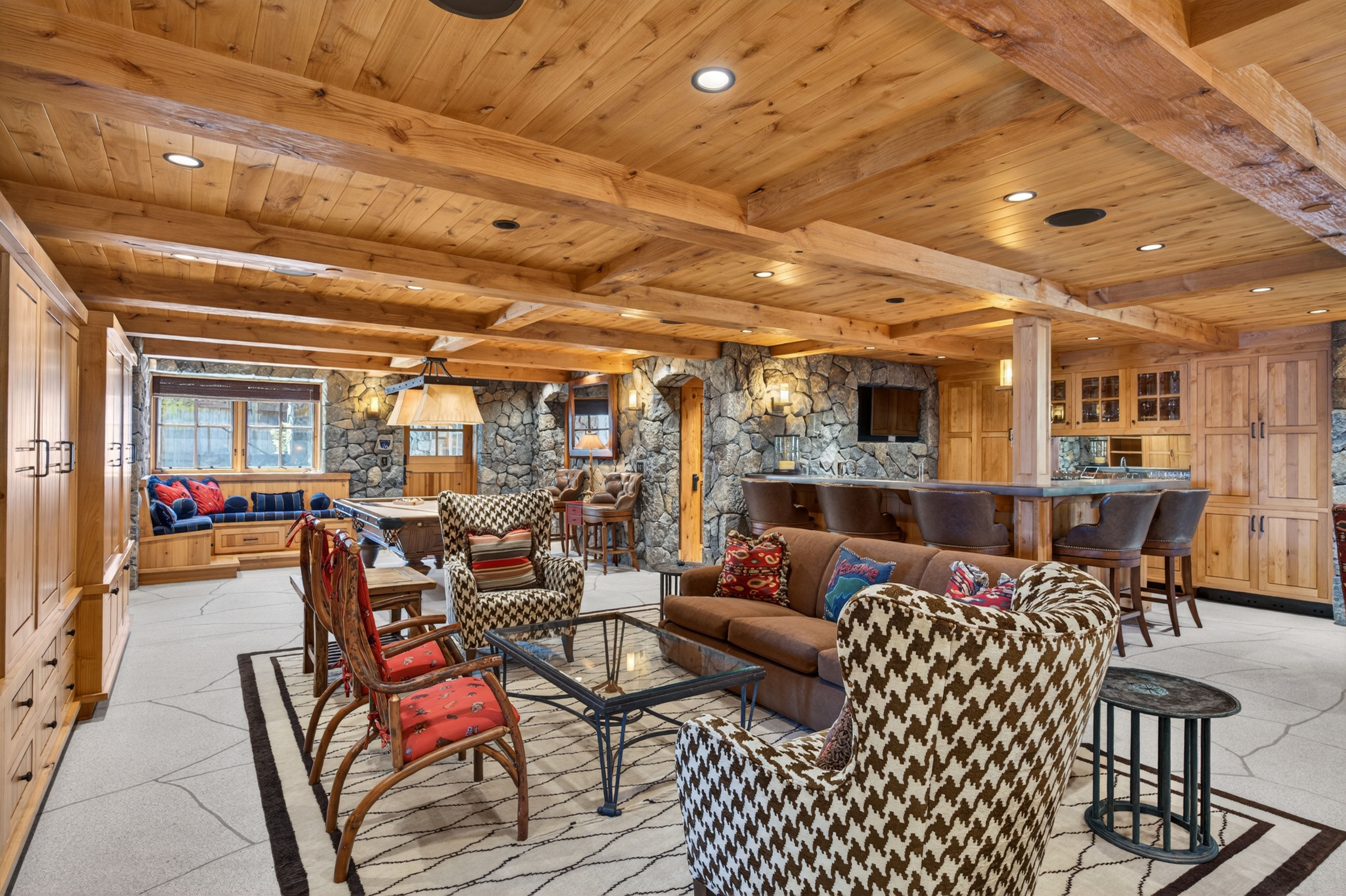 This expansive, rustic-style basement features stone accent walls, rich wood-beamed ceilings, and a high-end integrated bar area. The versatile space includes a living area with comfortable seating, a professional-style pool table, and cozy window bench seating, making it an ideal entertainment hub. The combination of high-quality wood cabinetry and natural stone creates a warm, mountain-lodge aesthetic throughout the room.