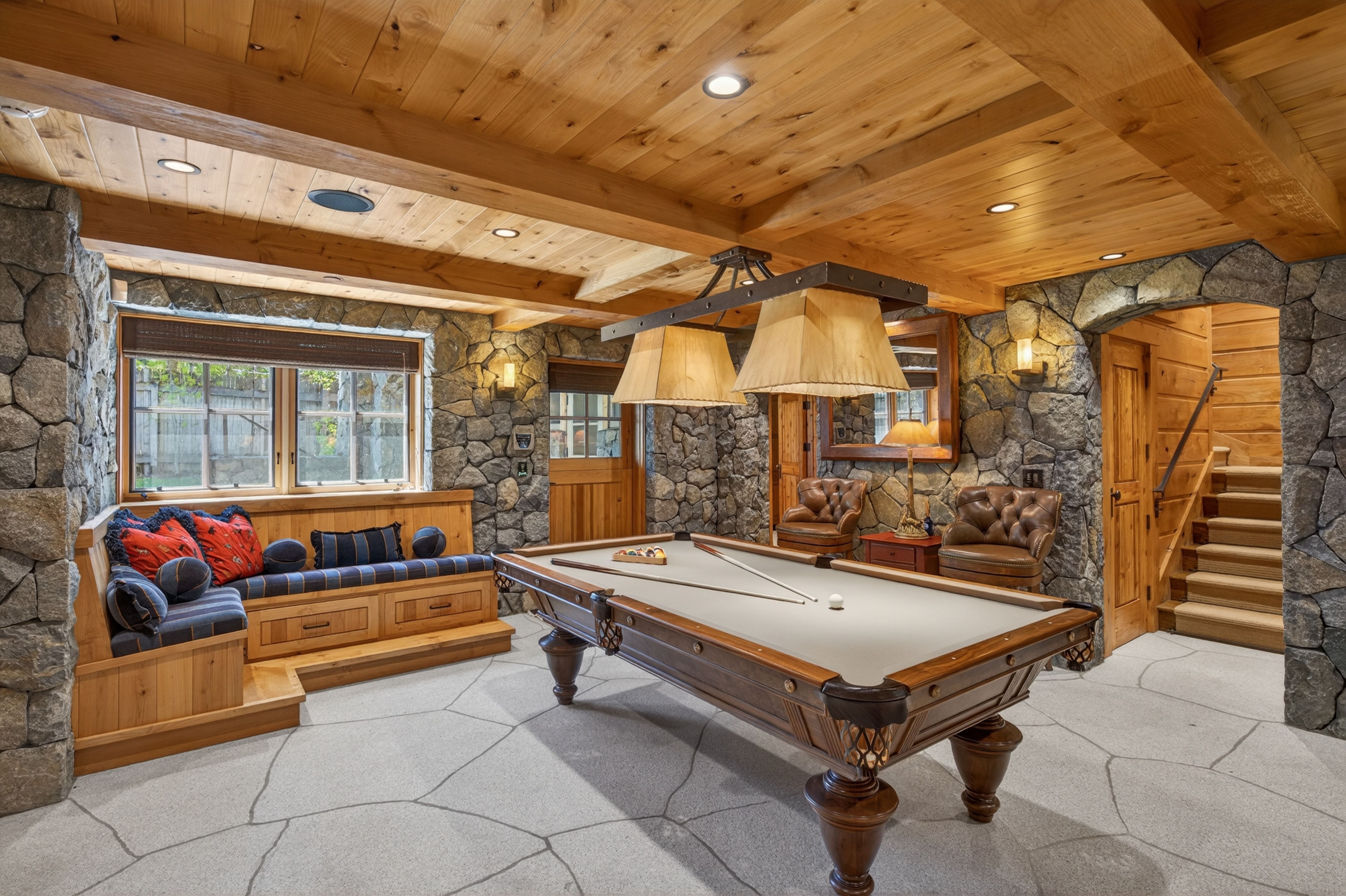 This rustic game room features a classic billiard table as the center stage, complemented by matching upholstered built-in seating and leather armchairs. The space showcases high-end finishes, including natural stone wall cladding, extensive wood paneling on the ceiling, and elegant stone flooring. It delivers a warm, lodge-like aesthetic that is both luxurious and inviting.