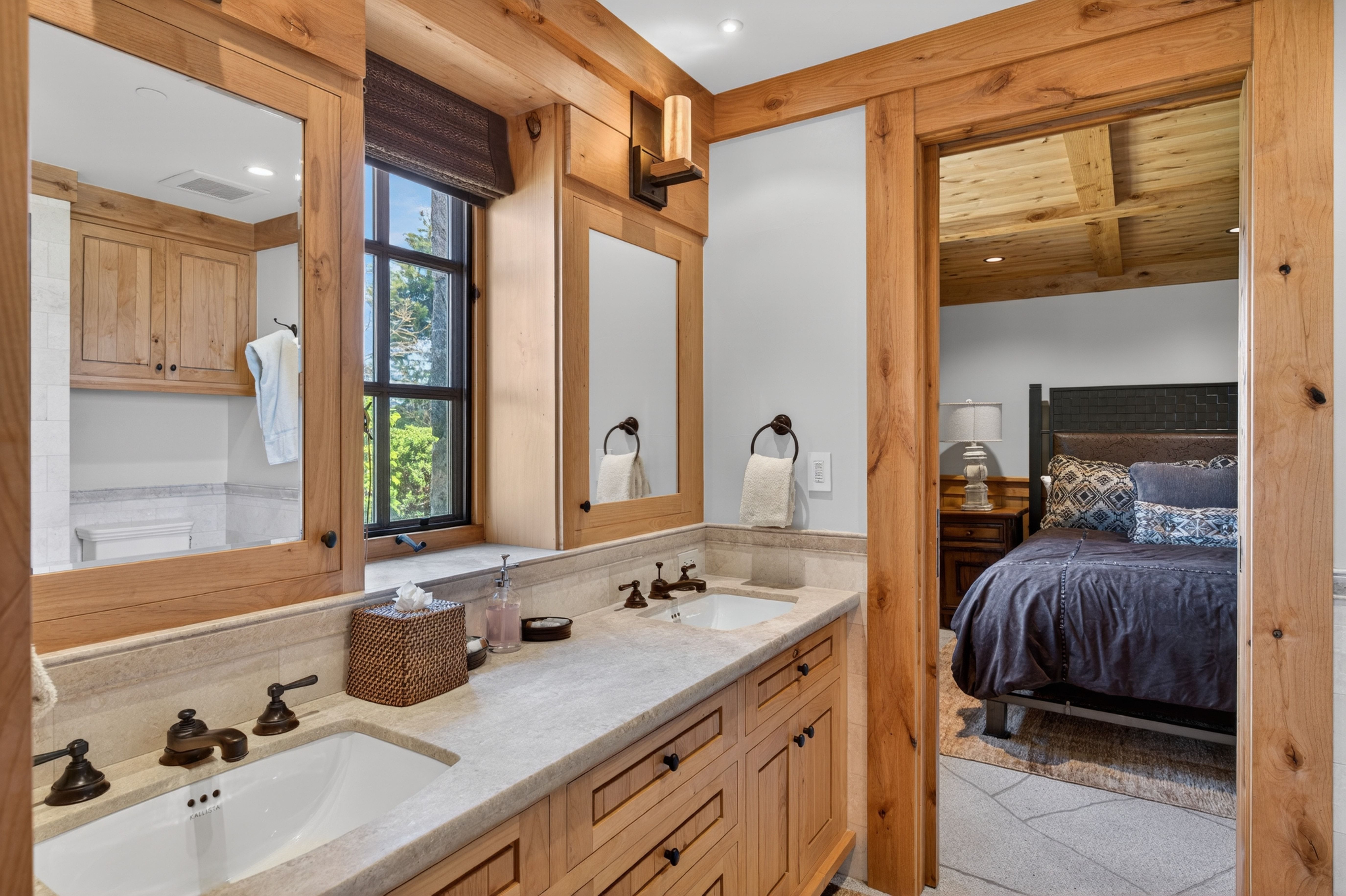 This primary bathroom features warm-toned wood cabinetry and neutral stone countertops, lending a high-end, rustic cabin aesthetic. The space includes dual vanity sinks, bespoke wood-trimmed mirrors, and direct access to an adjacent bedroom, creating a seamless and functional layout. The warm lighting and natural wood accents contribute to a luxurious yet cozy retreat atmosphere.