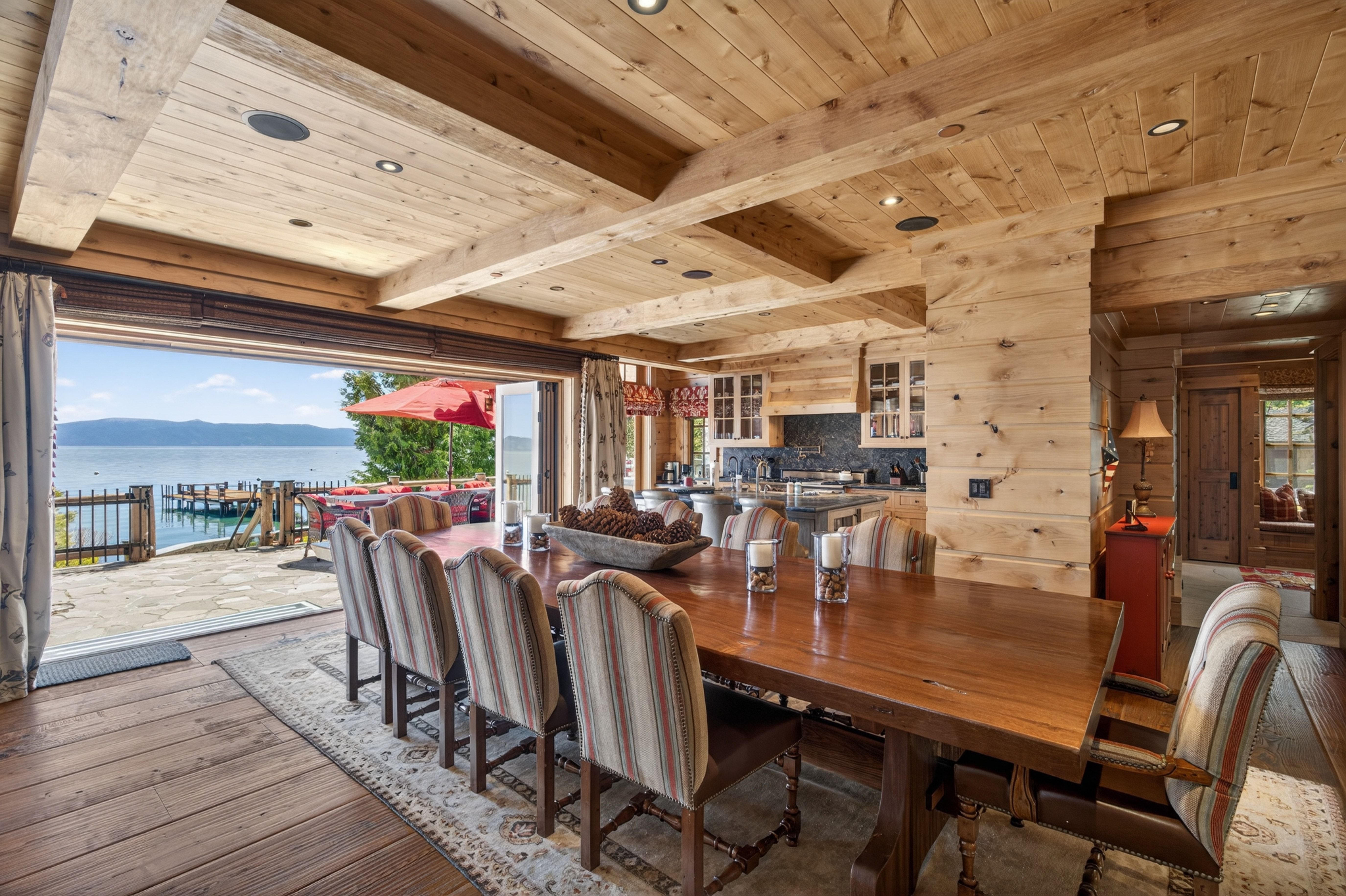 This grand dining area features a large, rustic wooden table bordered by striped upholstered chairs, set within a cozy, timber-clad lodge-style space. The room seamlessly connects to a stone-paved outdoor terrace through wide, open glass walls, offering picturesque views of the lake and dock. Natural wood textures throughout the ceiling and walls create an inviting, cabin-inspired atmosphere perfect for hosting.