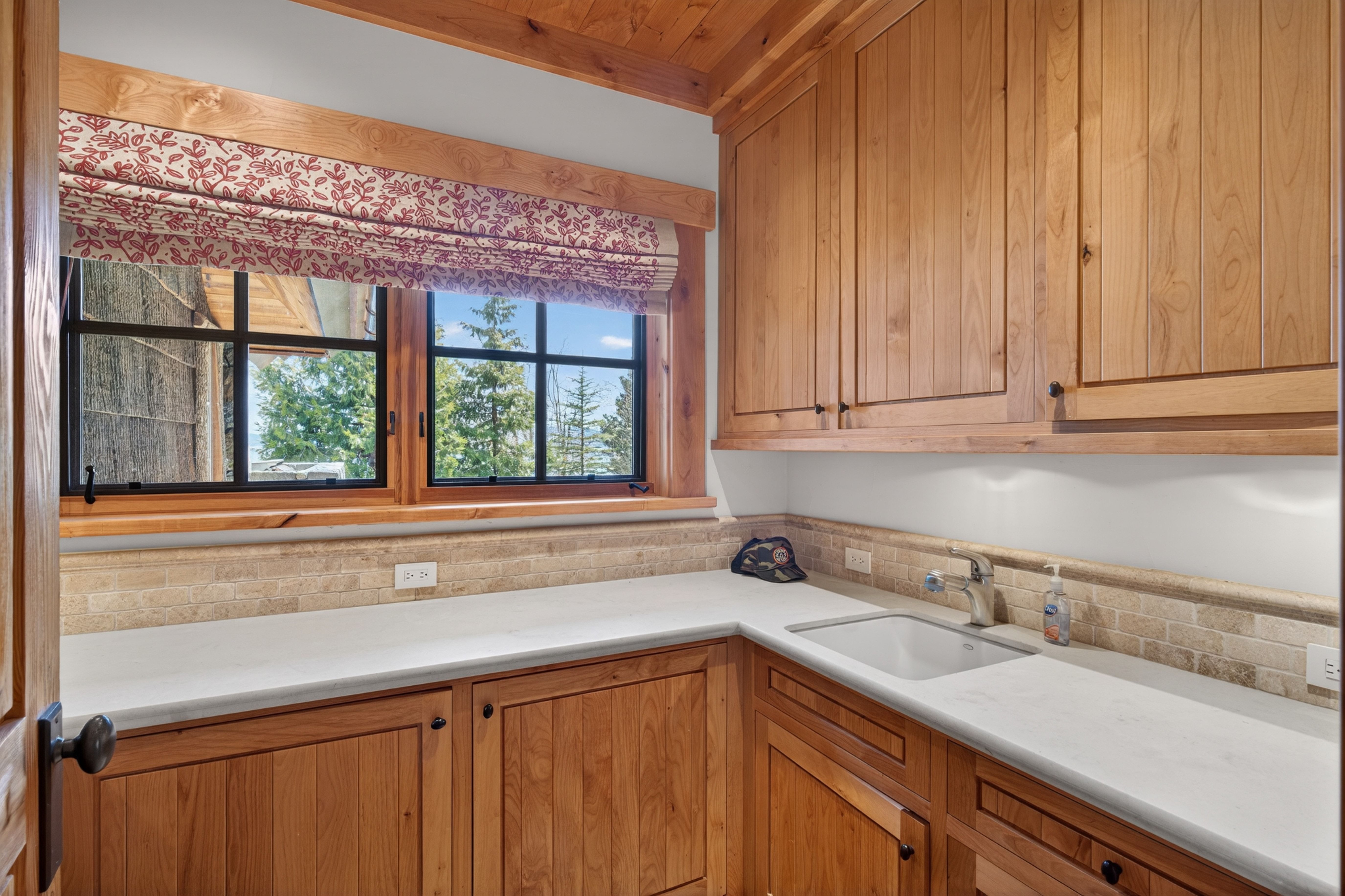 This interior image showcases a rustic-style kitchen space with rich, natural wood cabinetry and matching trim. The room features a white quartz countertop, a farmhouse-style sink, and a light-toned stone tile backsplash under a large multi-paned window adorned with a patterned valance. The area is well-lit and integrates warm, earthy materials to create a cozy and functional workspace.