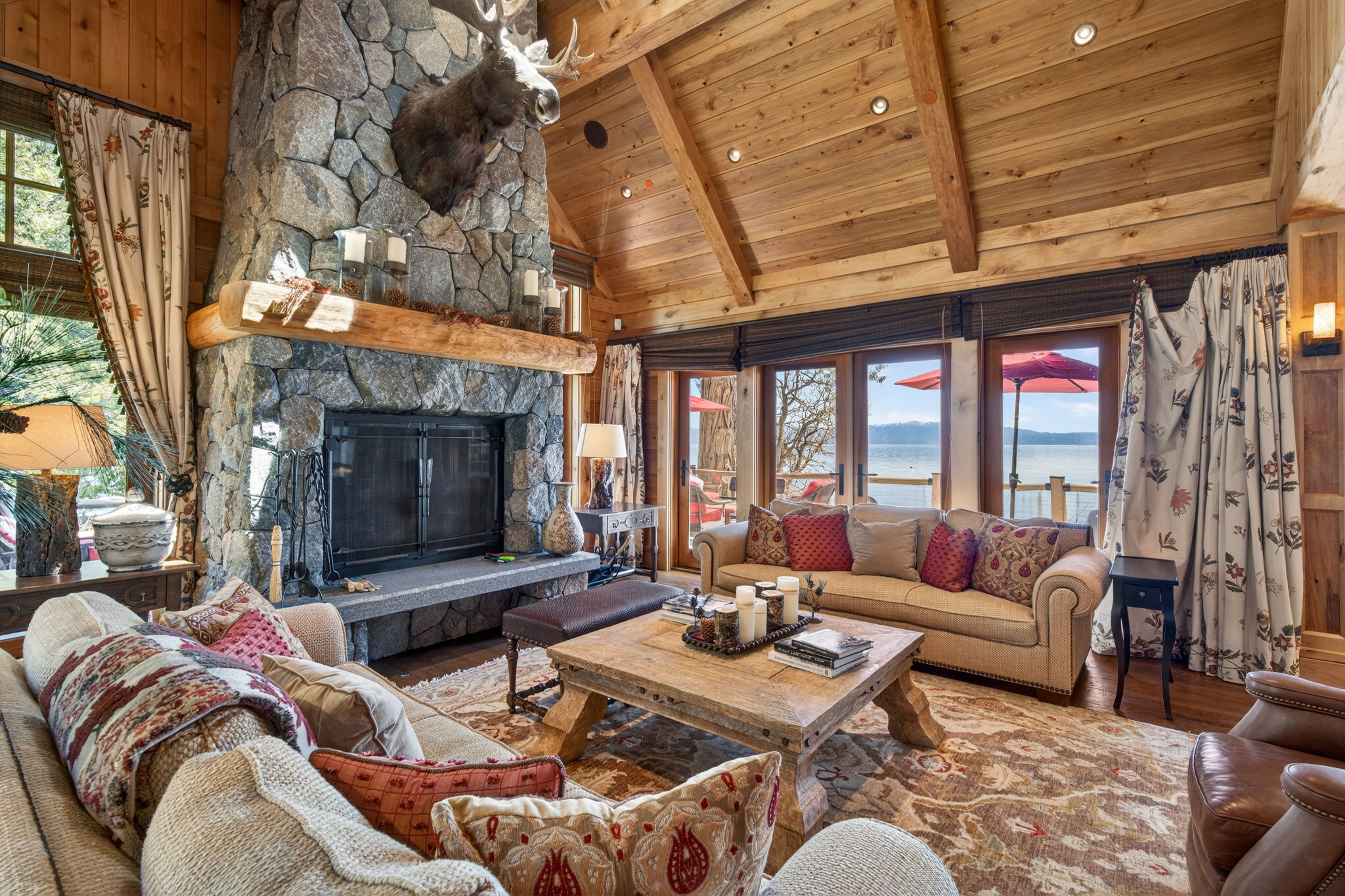 This cozy living room features a striking stone fireplace focal point adorned with a moose head mount and a heavy timber mantel. Large windows and glass doors offer stunning lake views, complemented by rustic wood-paneled, vaulted ceilings and rich, warm-toned furnishings. The space creates an inviting, upscale cabin aesthetic perfect for relaxing.